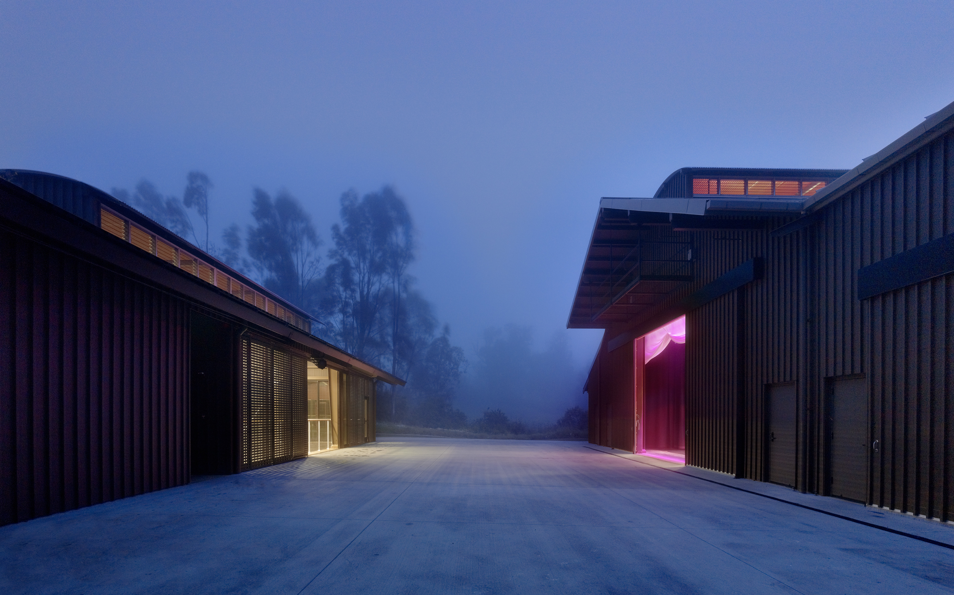 Exterior of two buildings with theater on the right with pink glow emitting and white yellow light coming from building on the left. Misty evening behind