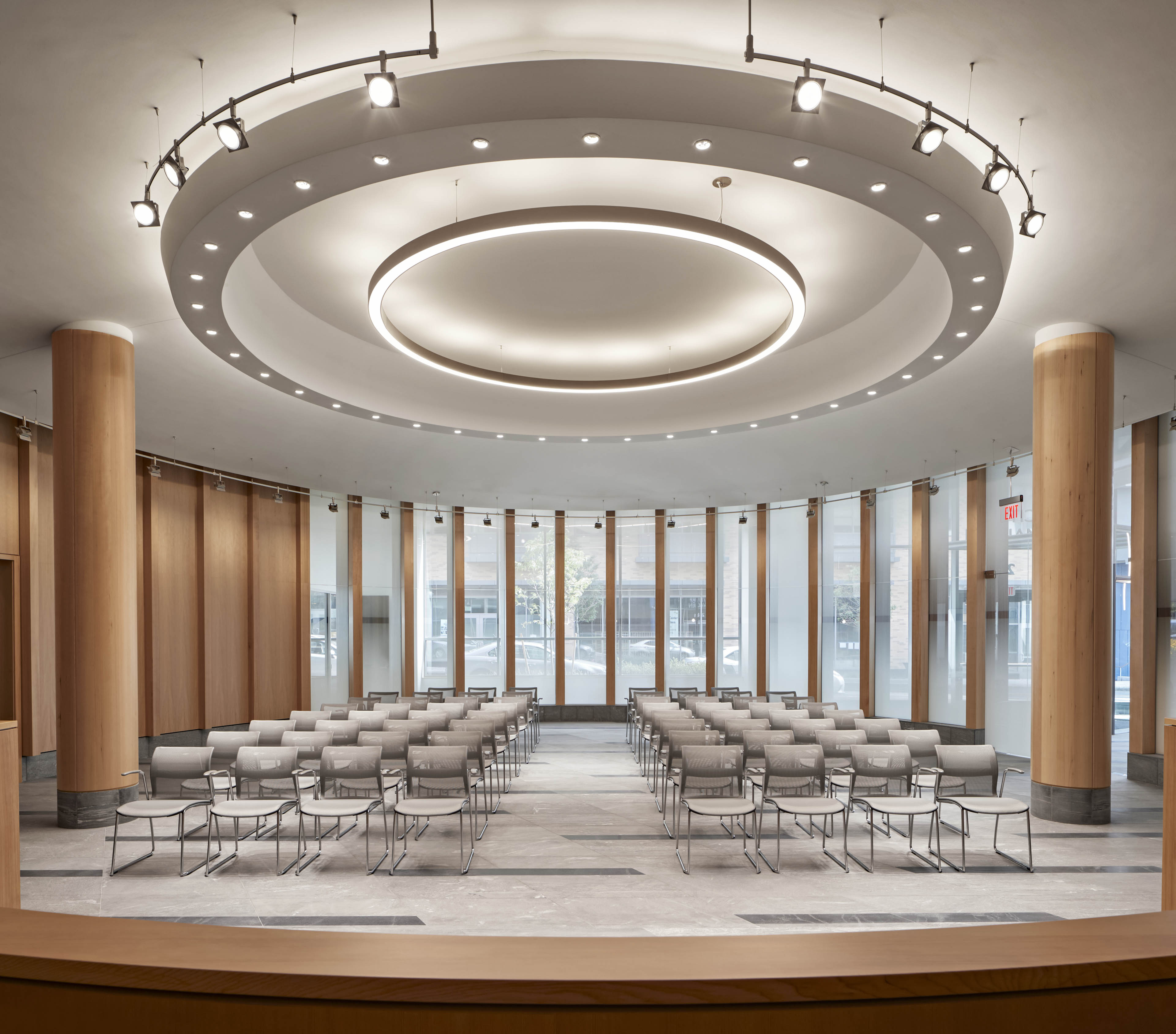Meeting room with large wood poles on left and right and alternating wood and tall vertical windows curving around wall with bright lights and seats in center