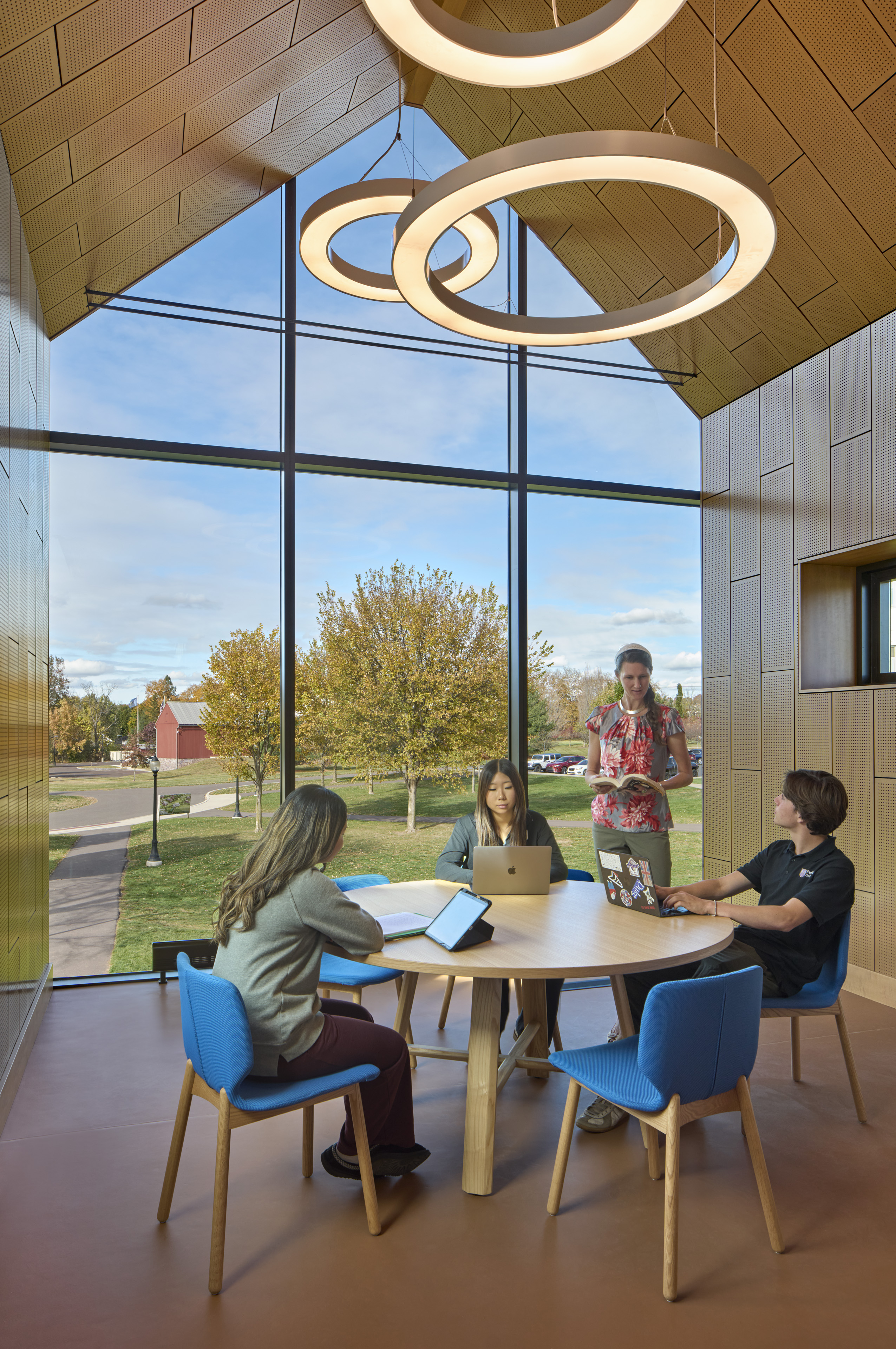  Students seated around table with windows behind looking out to field with trees. circular lights hang above