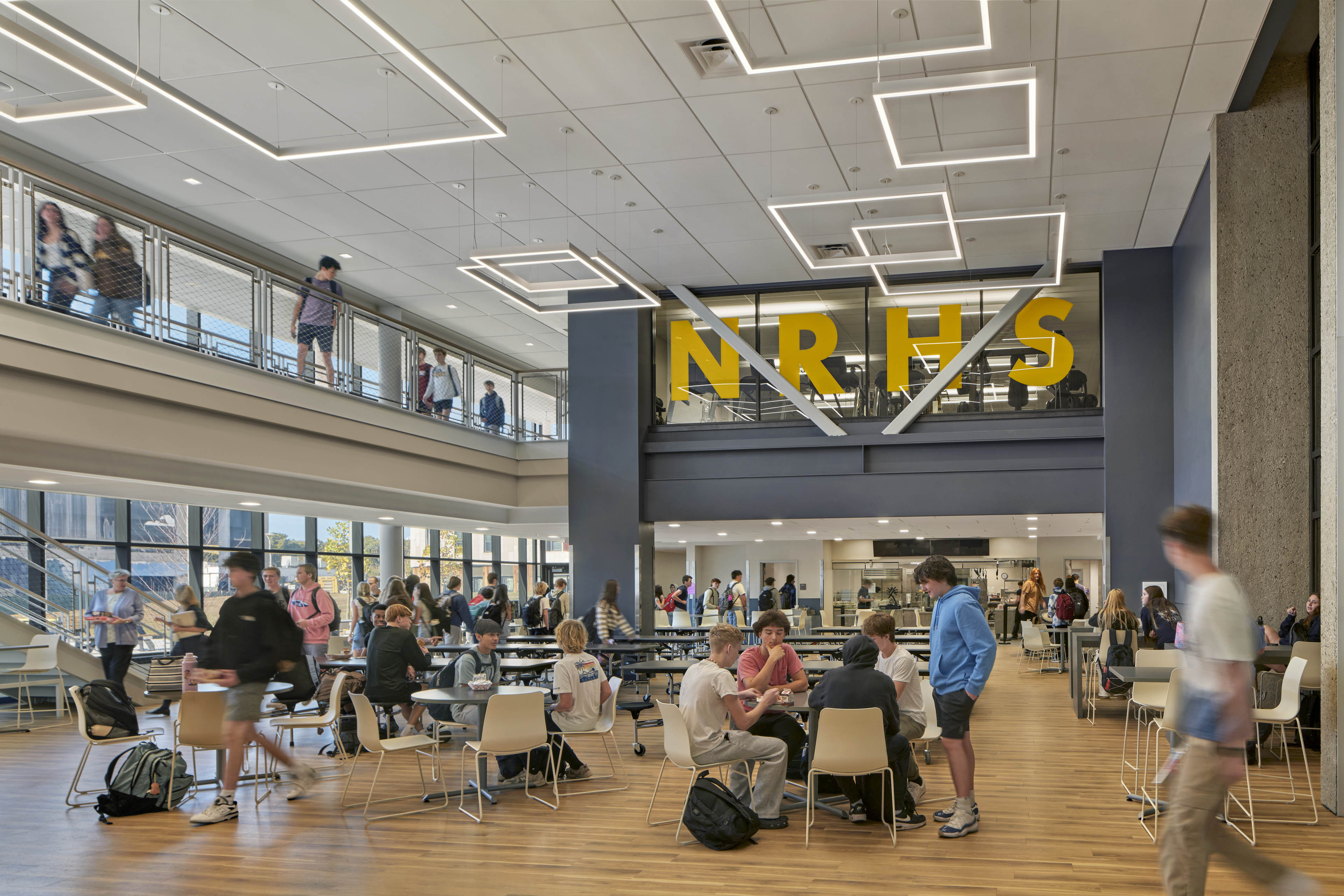 Students at cafeteria with clusters of tables and chairs, walkway on upper floor to left and school logo in yellow on windows above looking at gym
