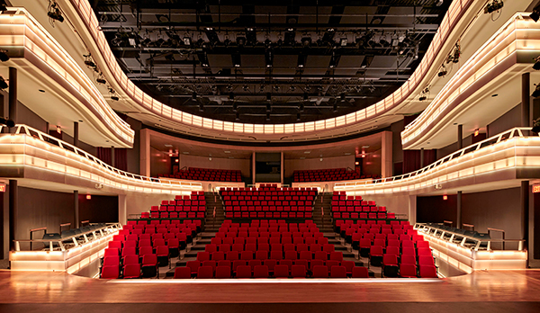 Theater with rounded seating area with red seats, 3 balconies illuminated from stage view