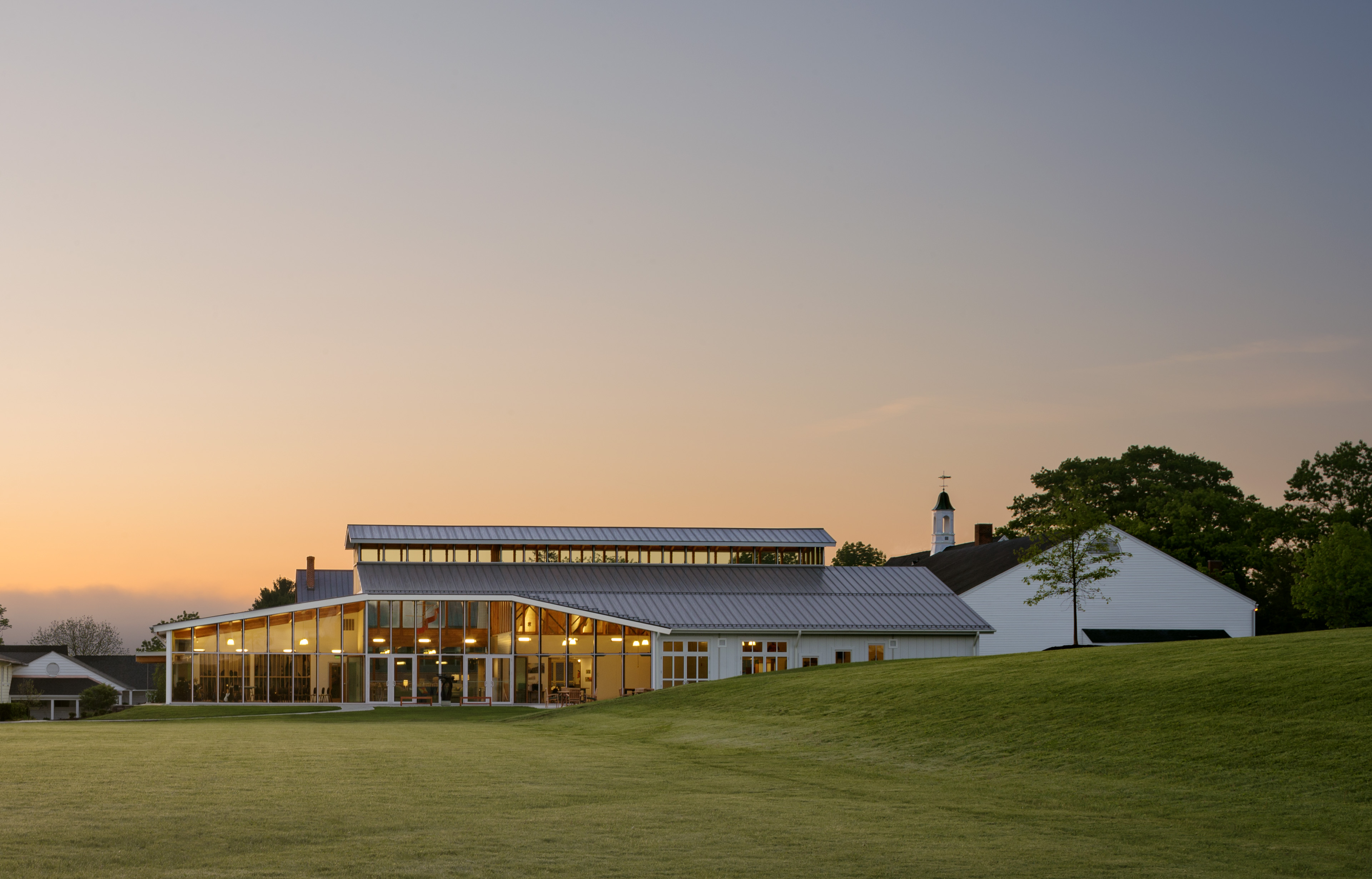 View of exterior at sunset with faint glow coming from school, rolling grass hill, and yellow orange sunset behind