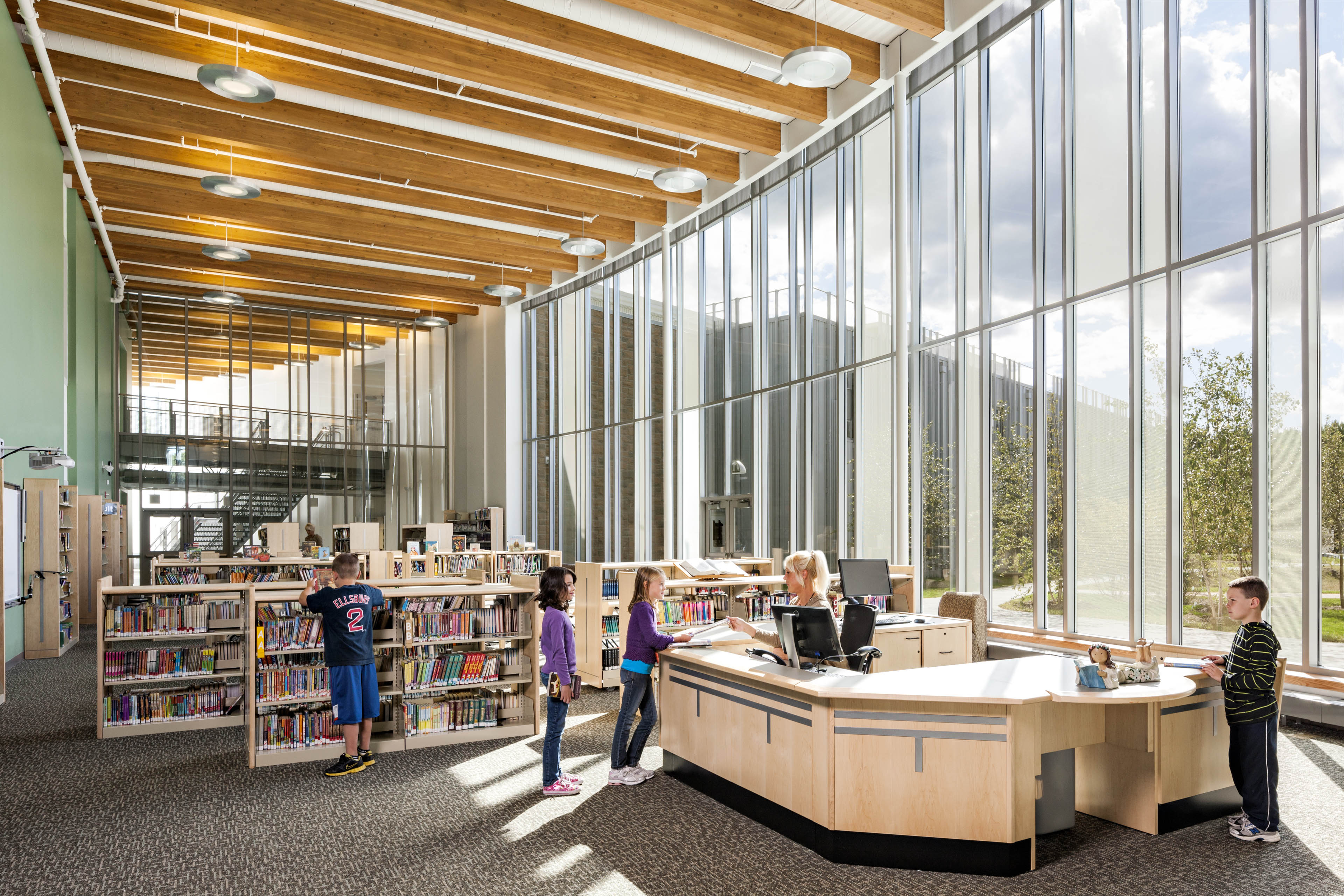 Students using library with sage green wall to the left and shelves of books in center with floor to ceiling windows on the right