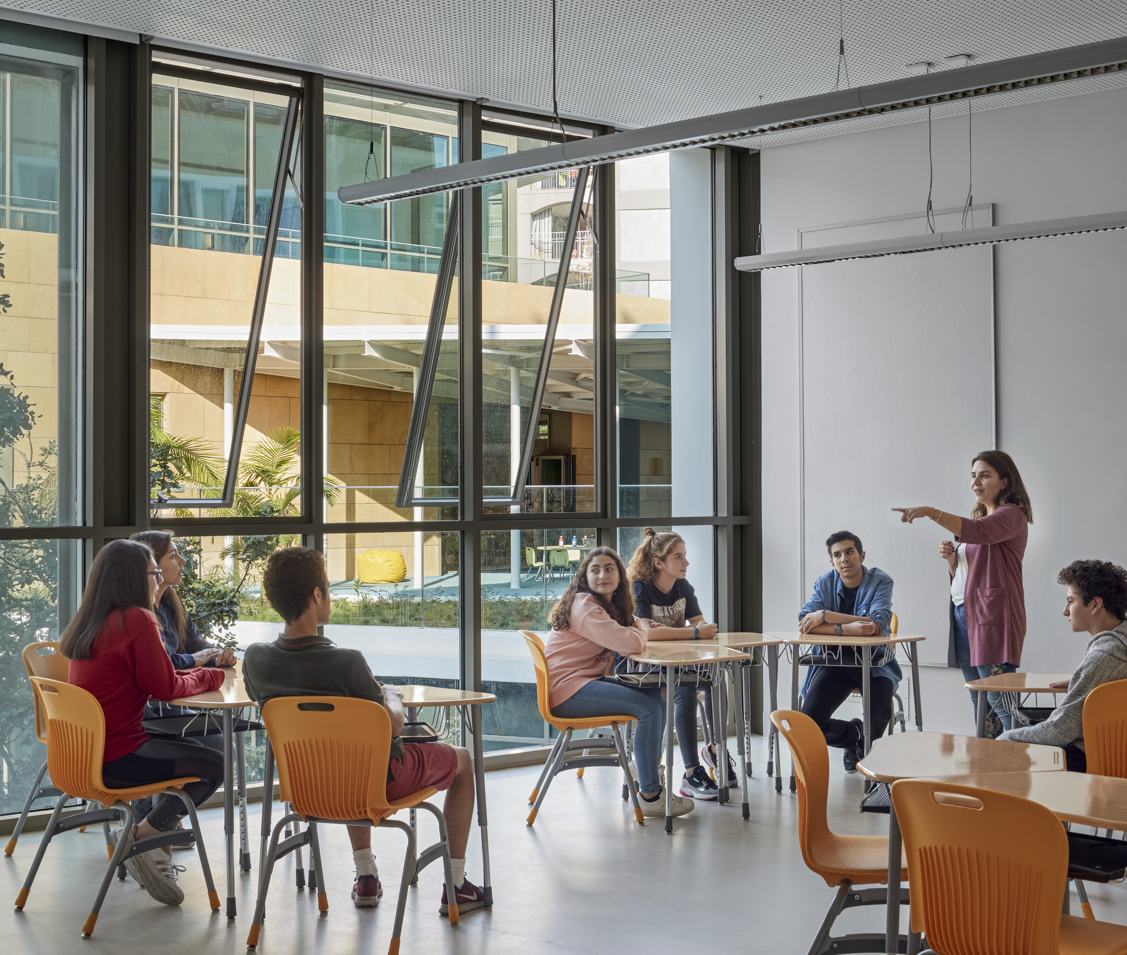 Students seated in classroom during instruction at tables with orange chairs and windows to the left looking out to courtyard