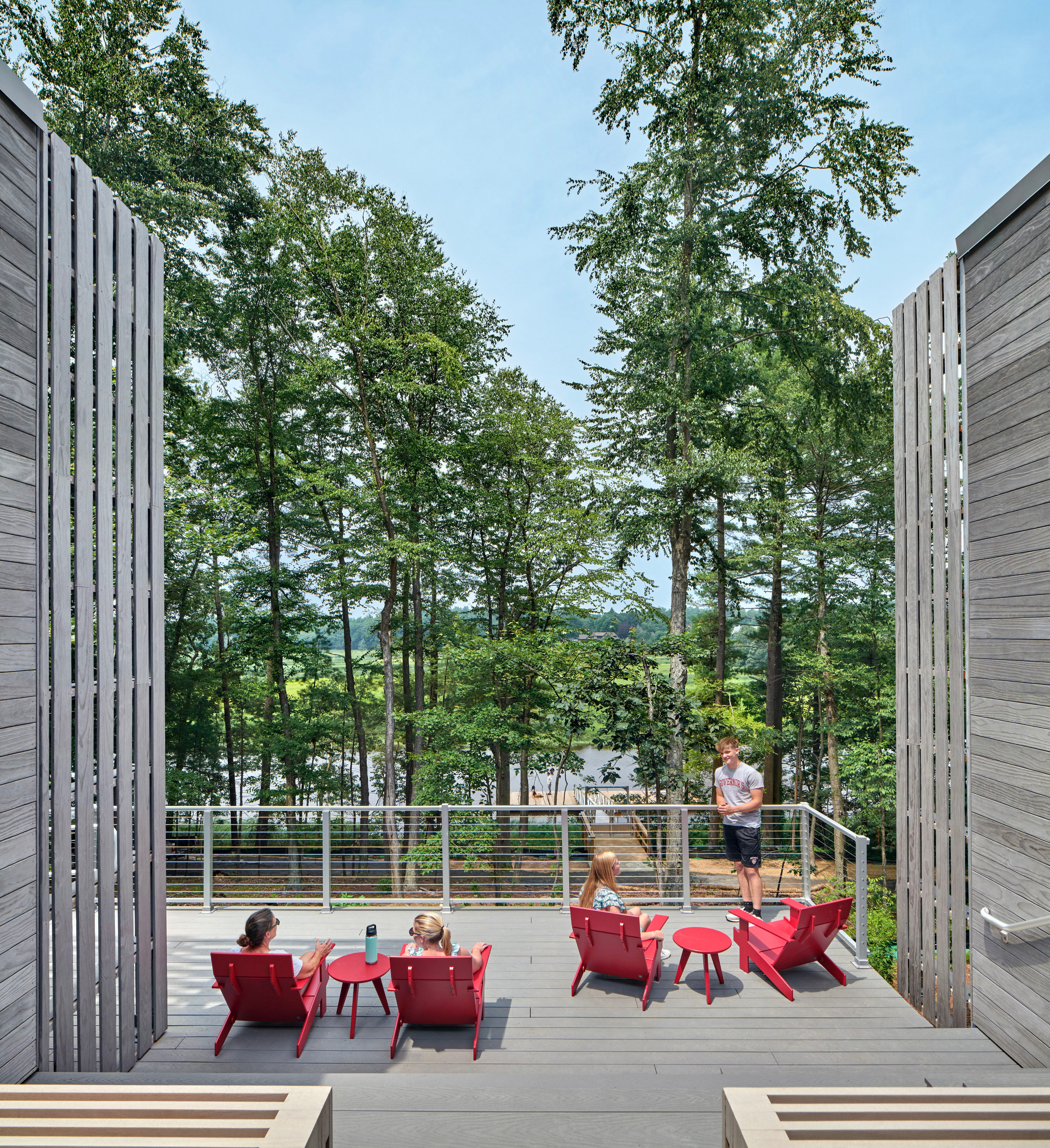 Students lounging on deck in red Adirondack chairs looking out at tall trees. Wood paneling on left and right