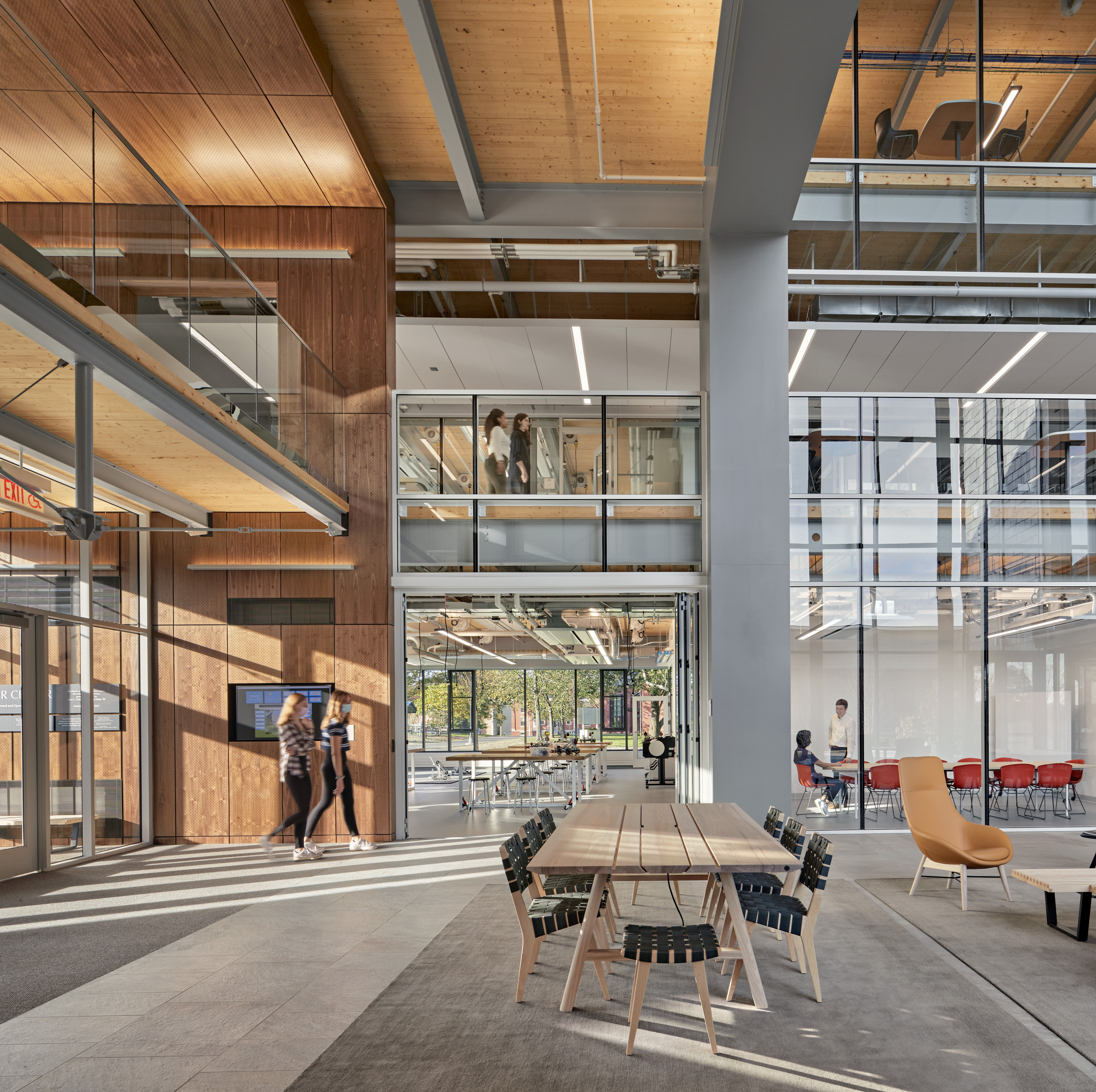 Common Area with long table and chairs, wood siding and paneling on ceiling to the left with students walking though with natural light. Classrooms with large windows in the back and to the right