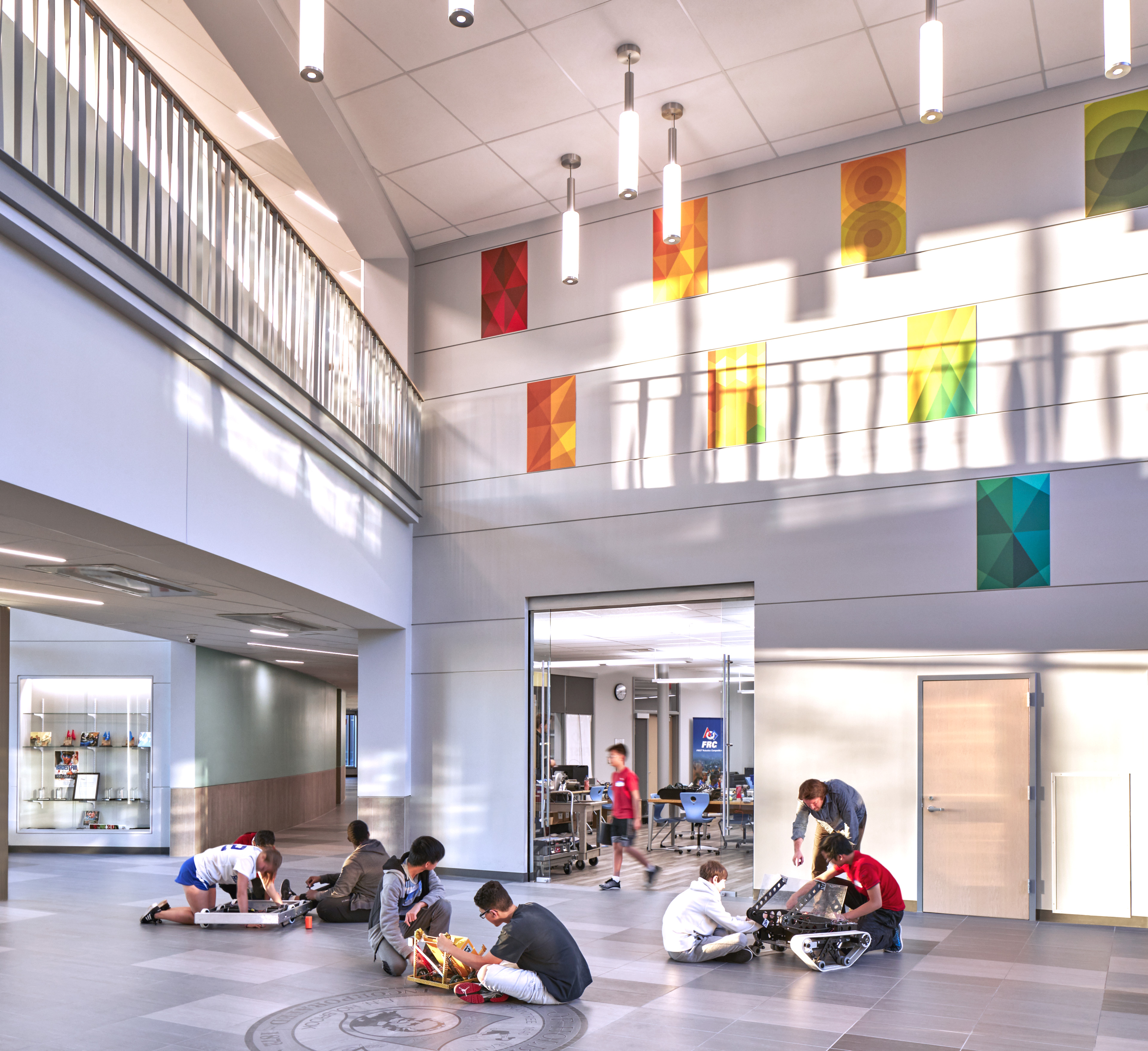 students seated on floor with robotics with classroom behind, yellow/orange artwork on second floor with light coming in