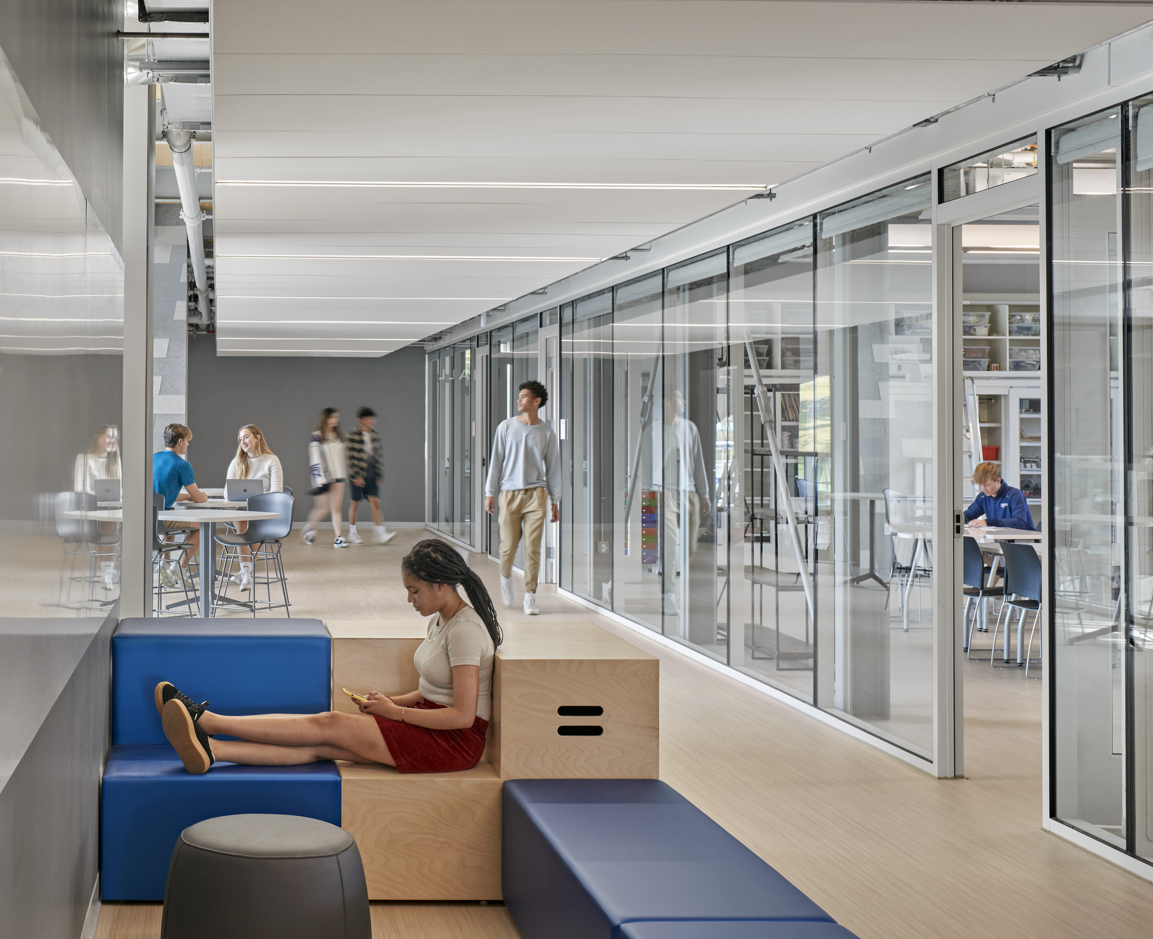 Hallway with lounge seating and students reading, working, and walking. Work rooms to the right with glass wall