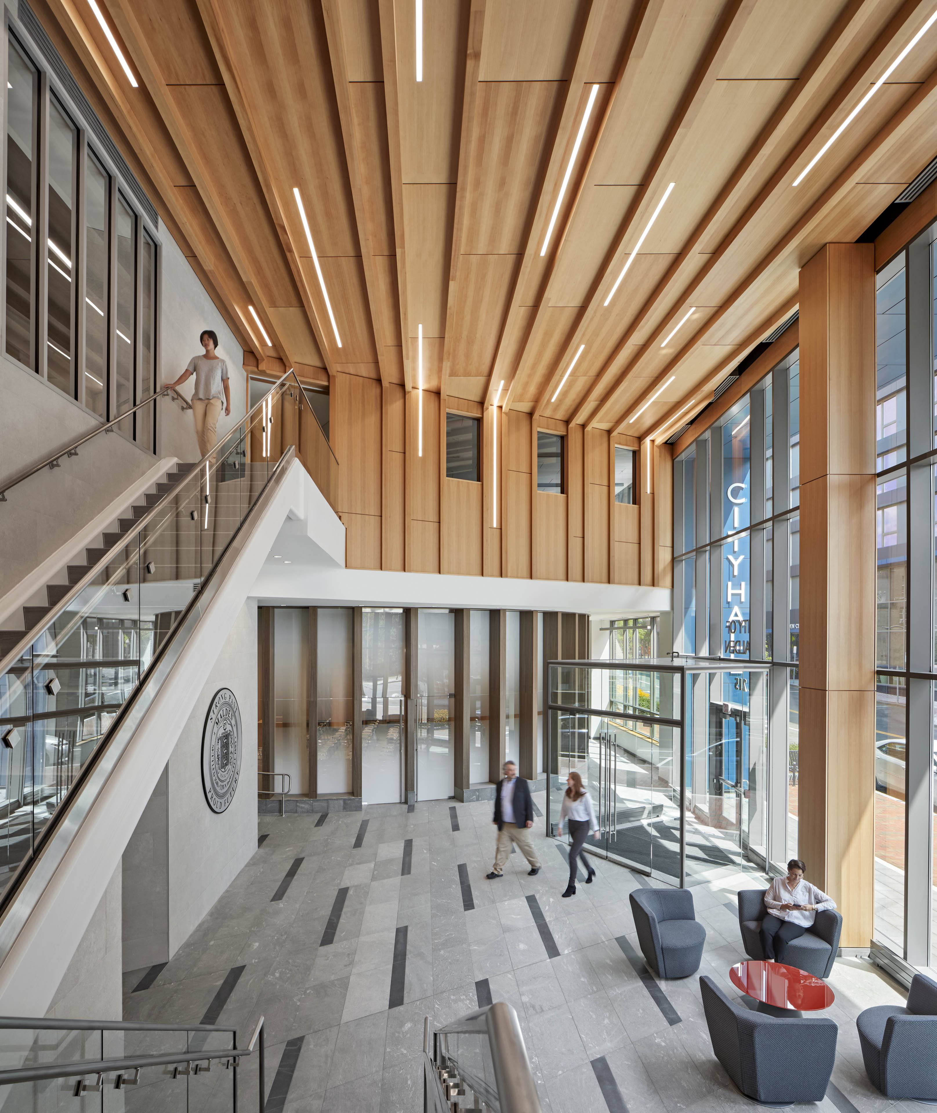 View from interior staircase looking at people walking through lobby with additional staircase to the left. Light wood starts on second floor and extends to ceiling