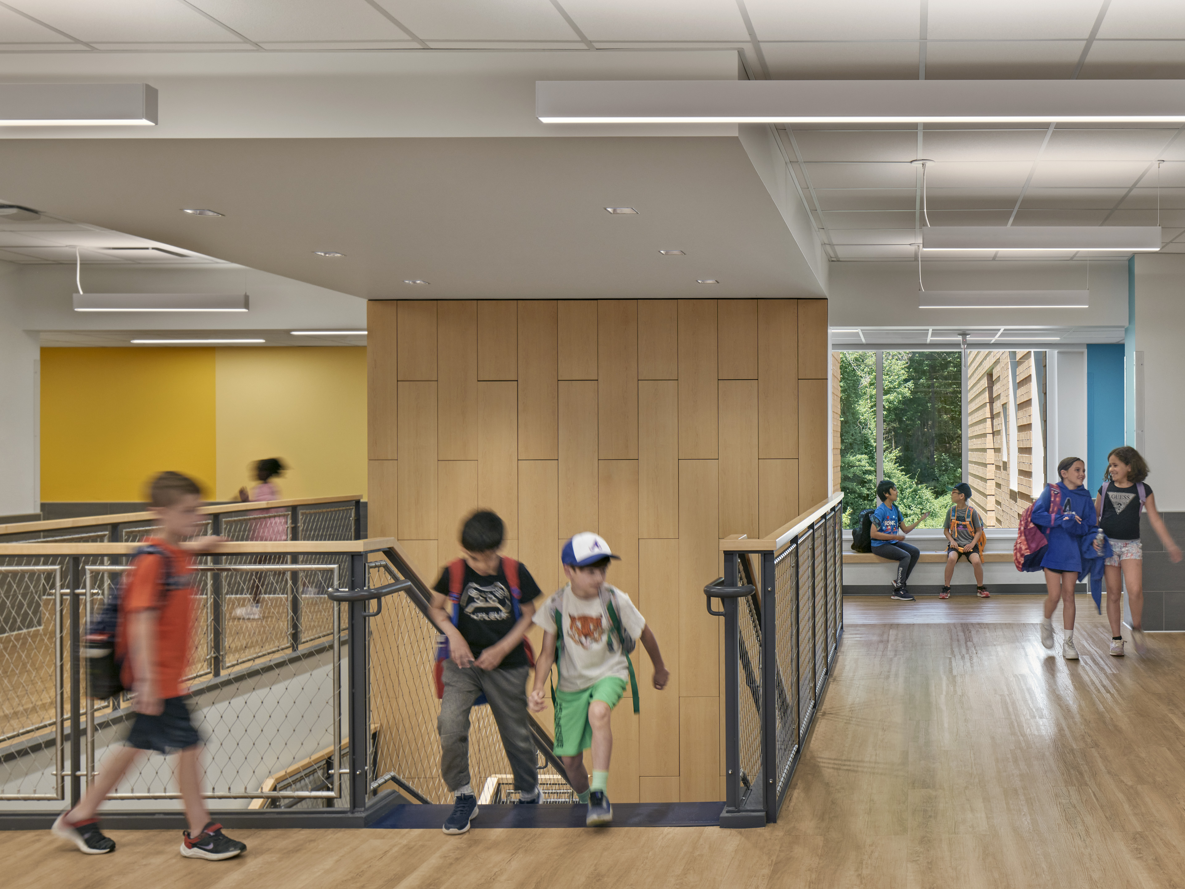 Students walking up stairs and using interior corridor to the right with window behind looking out to greenery