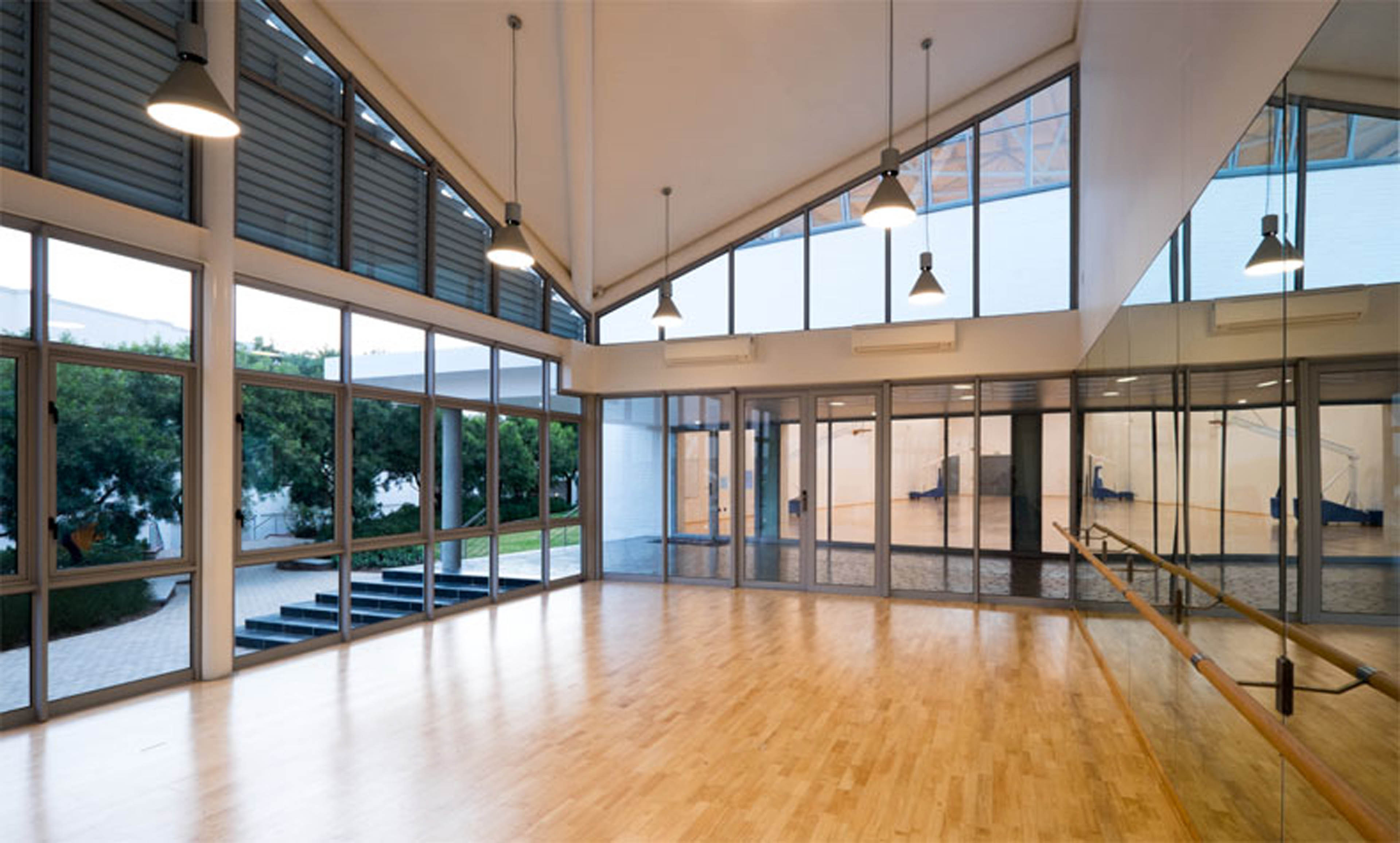 Dance room of gym with barre and mirror to right and windows looking out to the right. Large windows on top allowing natural light