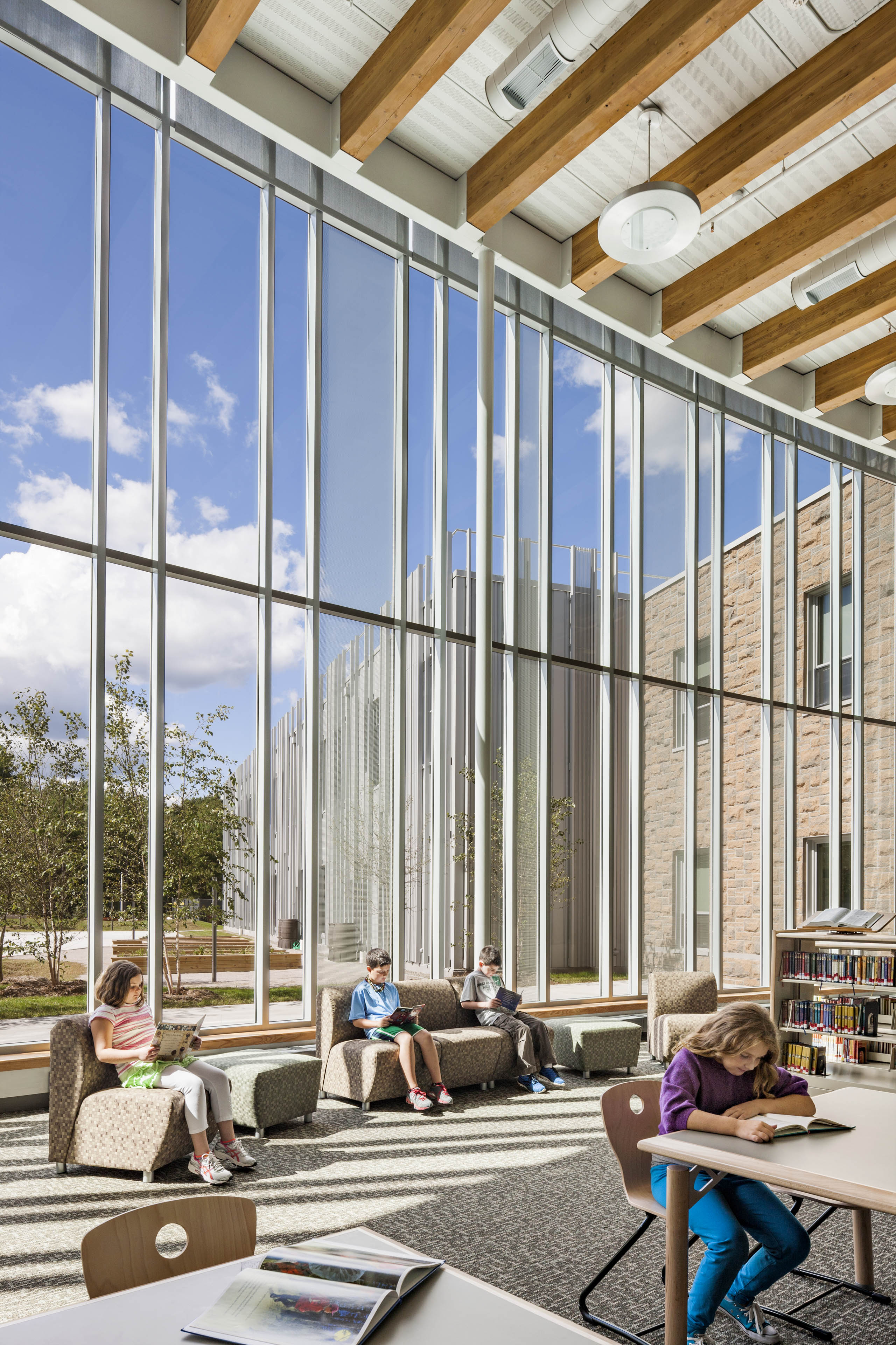 Students reading at lounge chairs in library with floor to ceiling walls on left side looking out to courtyard and books shelves to the right