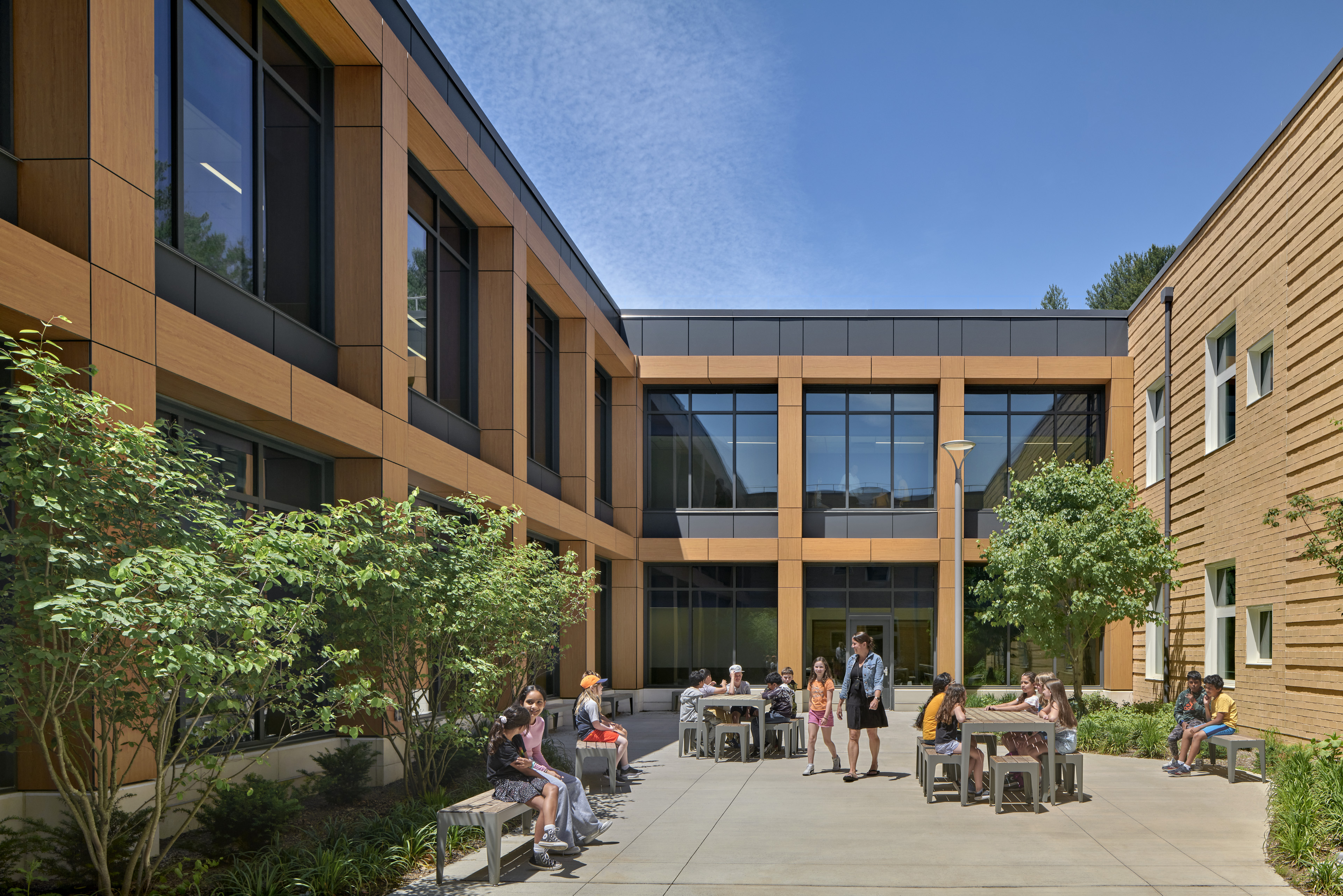 Interior courtyard of David Mindess School in Ashland, MA, students sat at long communal tables/benches. Building is light brown with black accents with sets of 3 vertical windows in a square.  