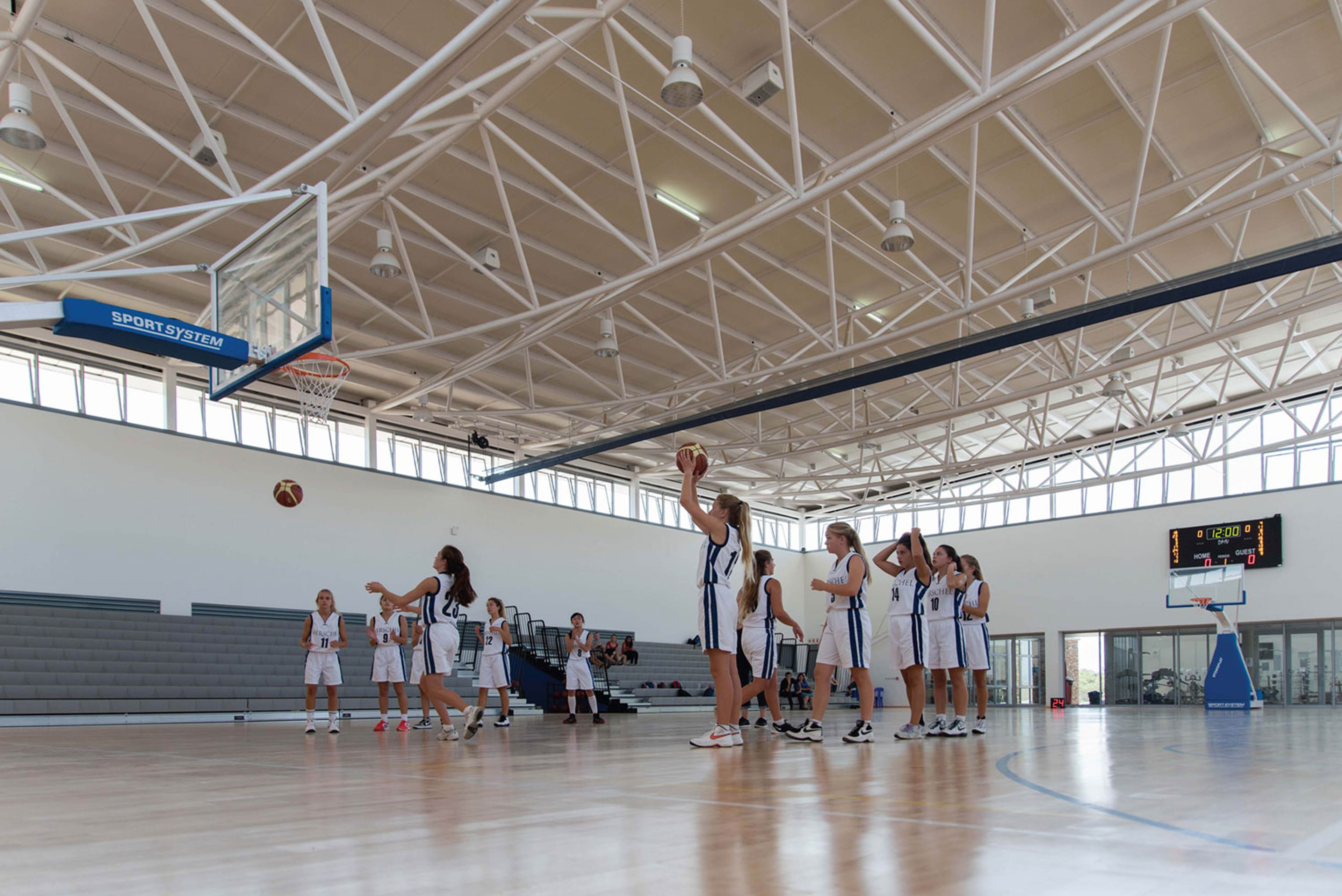 Gymnasium with basketball court and players practicing, hoop to the left and stadium seating behind