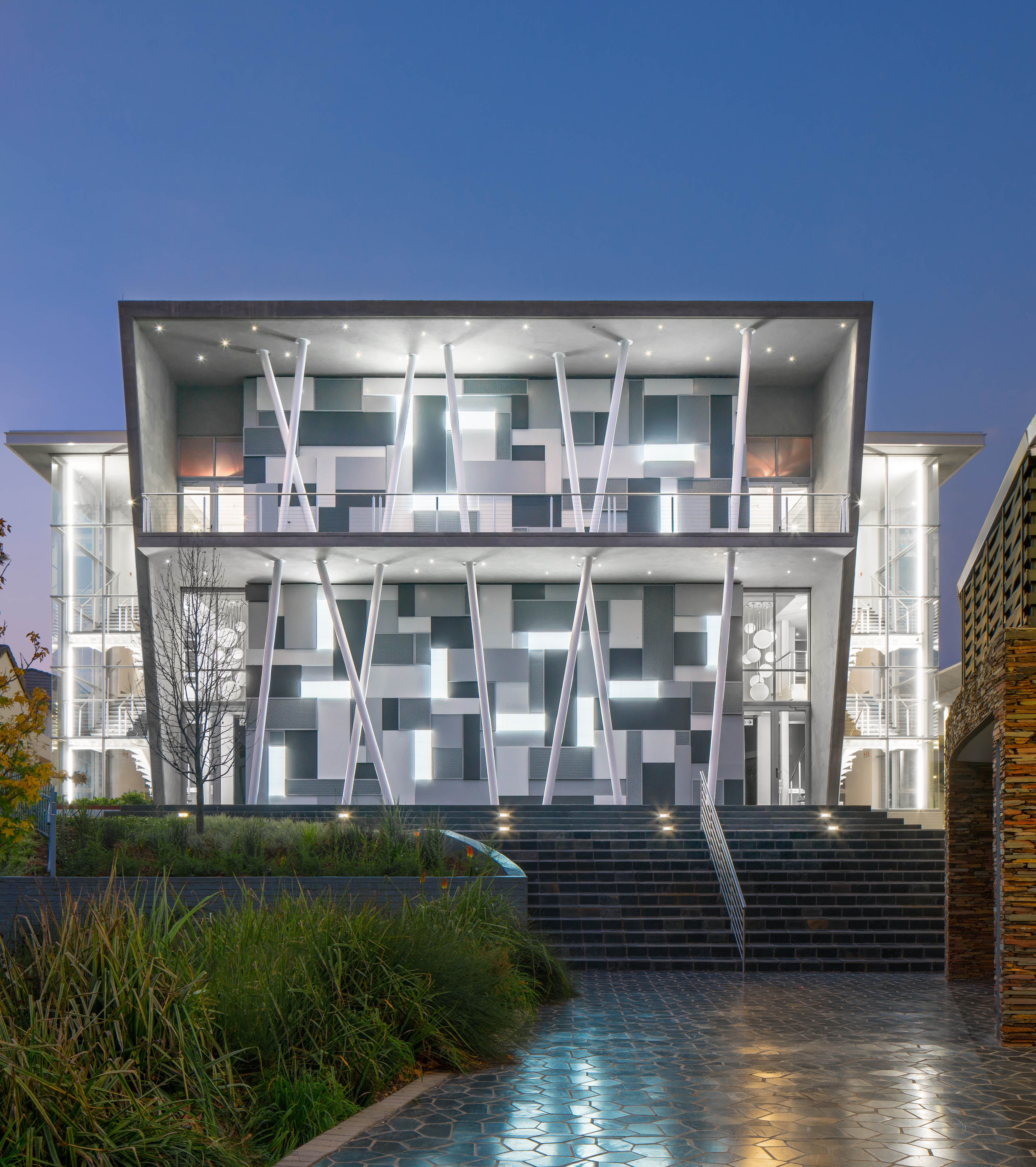 Front entrance of building at dusk, with stairs leading up and poles criss-crossing in front. Building behind lit up and extending past on left and right side