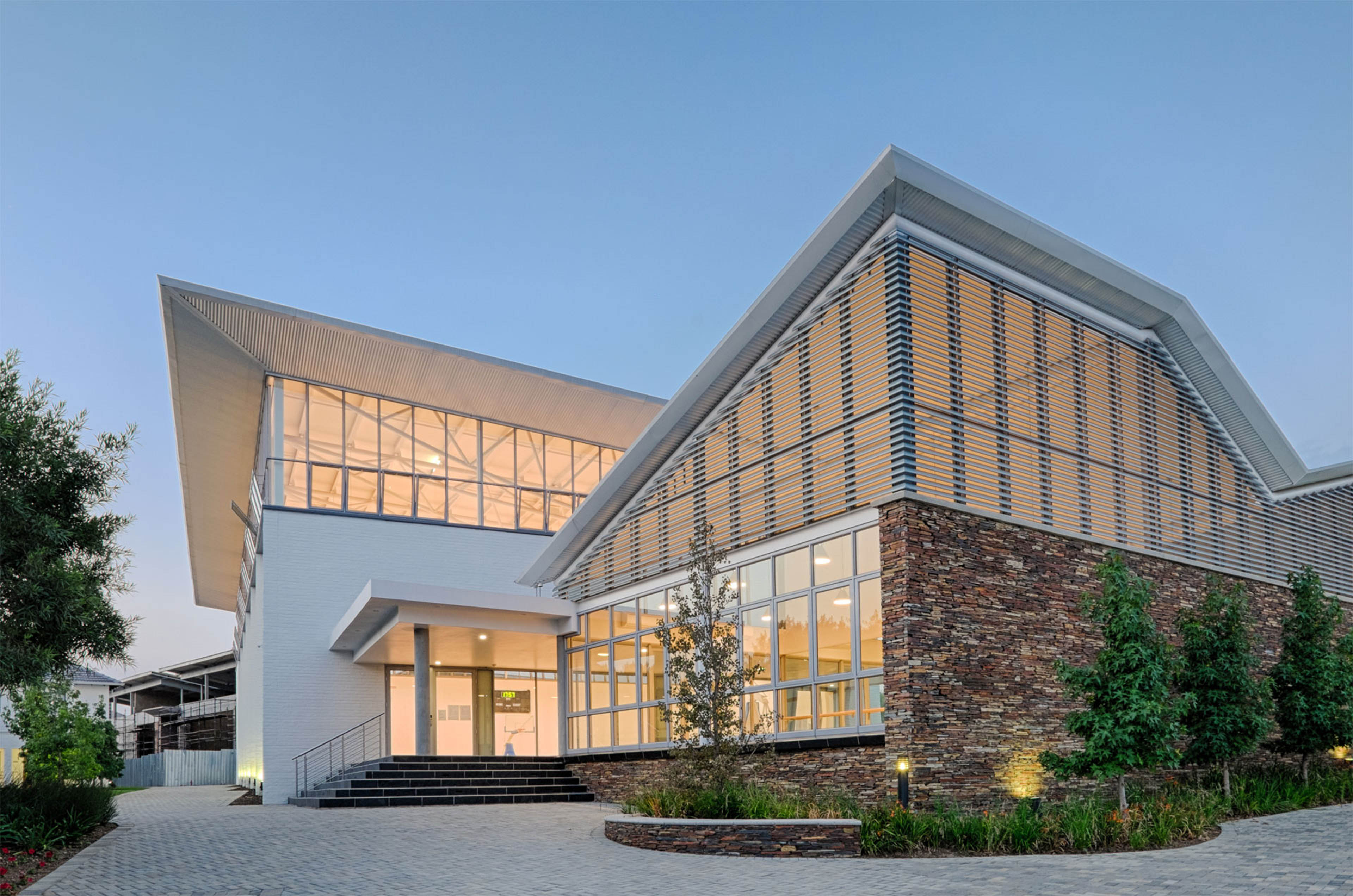 Exterior of building at dawn from side angle, yellow light emitting from windows placed all throughout. Small trees lining walkway to left and right