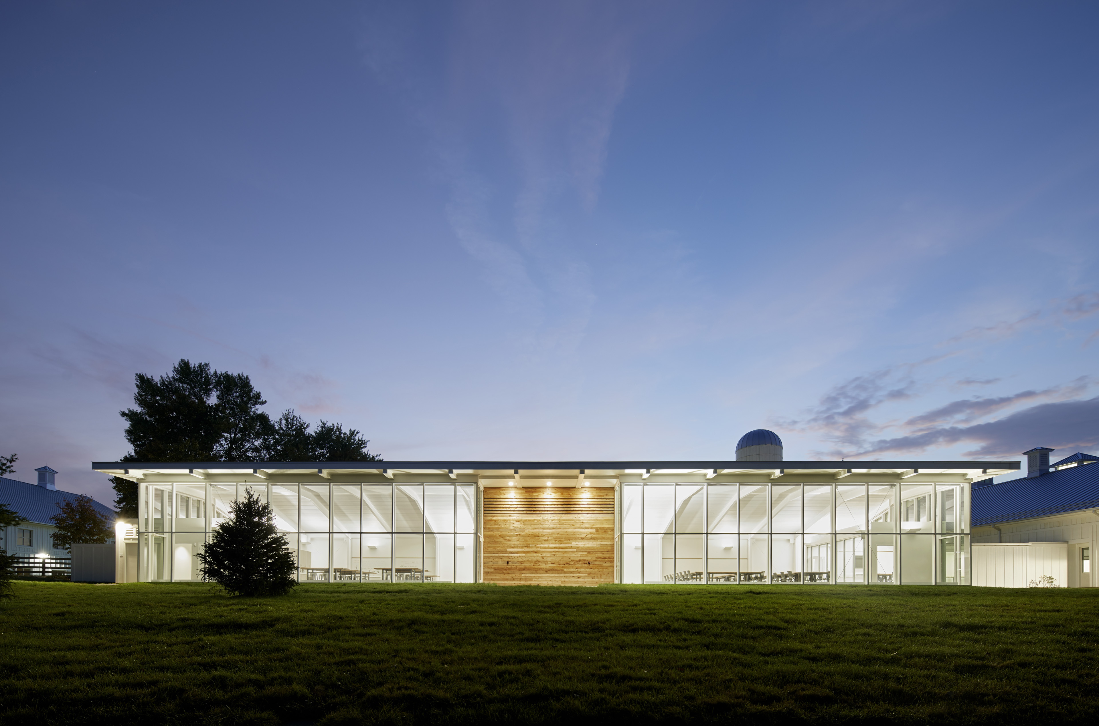 View of school at night, floor to ceiling vertical windows to left and right, light colored wood in center. Dusk sky behind and bight light emitting from building