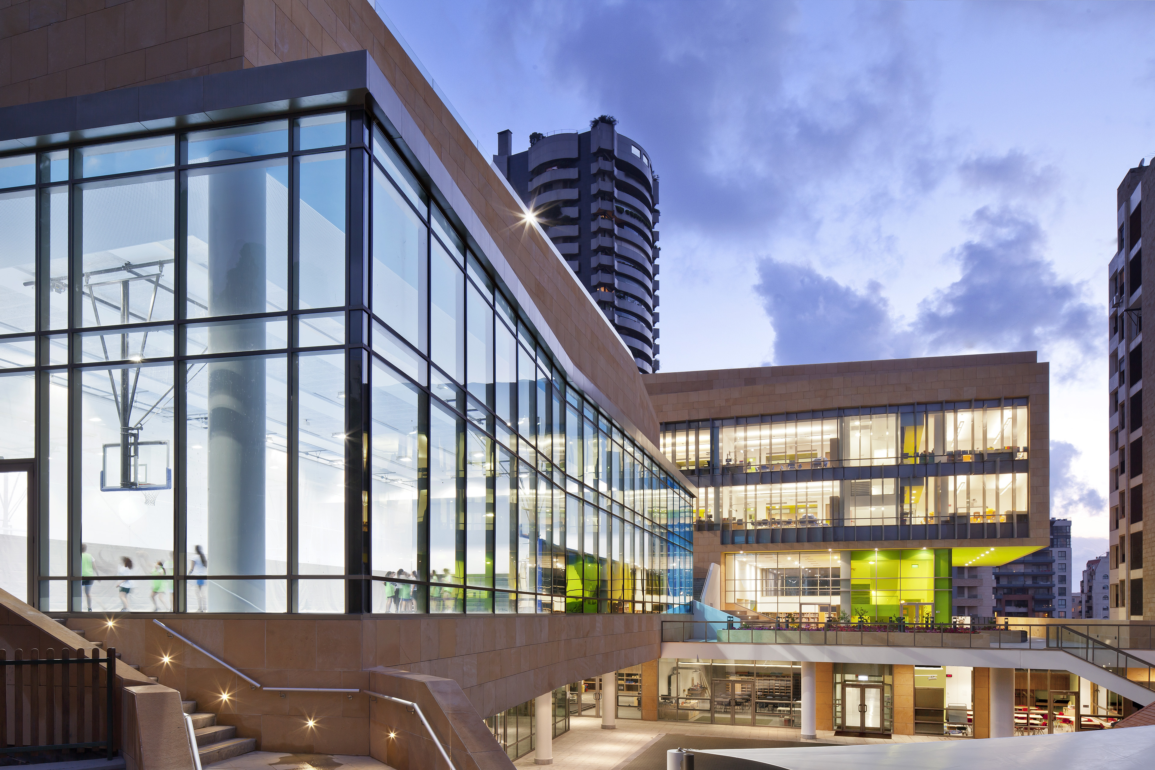 View of exterior at dusk with large vertical windows to the left with blue lights and windows in back with yellow light