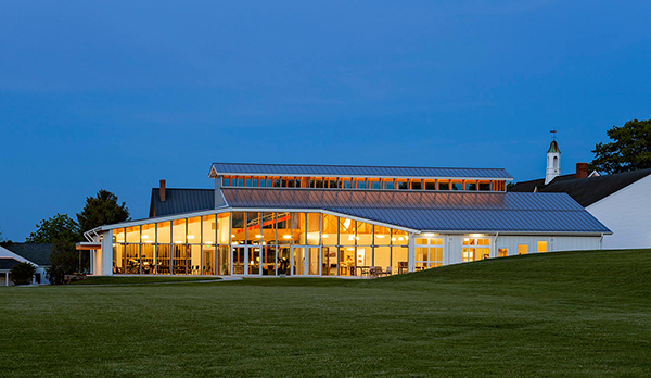 View at night of school from grassy hill, with long vertical windows with yellow orange light on building. Dark blue sky behind