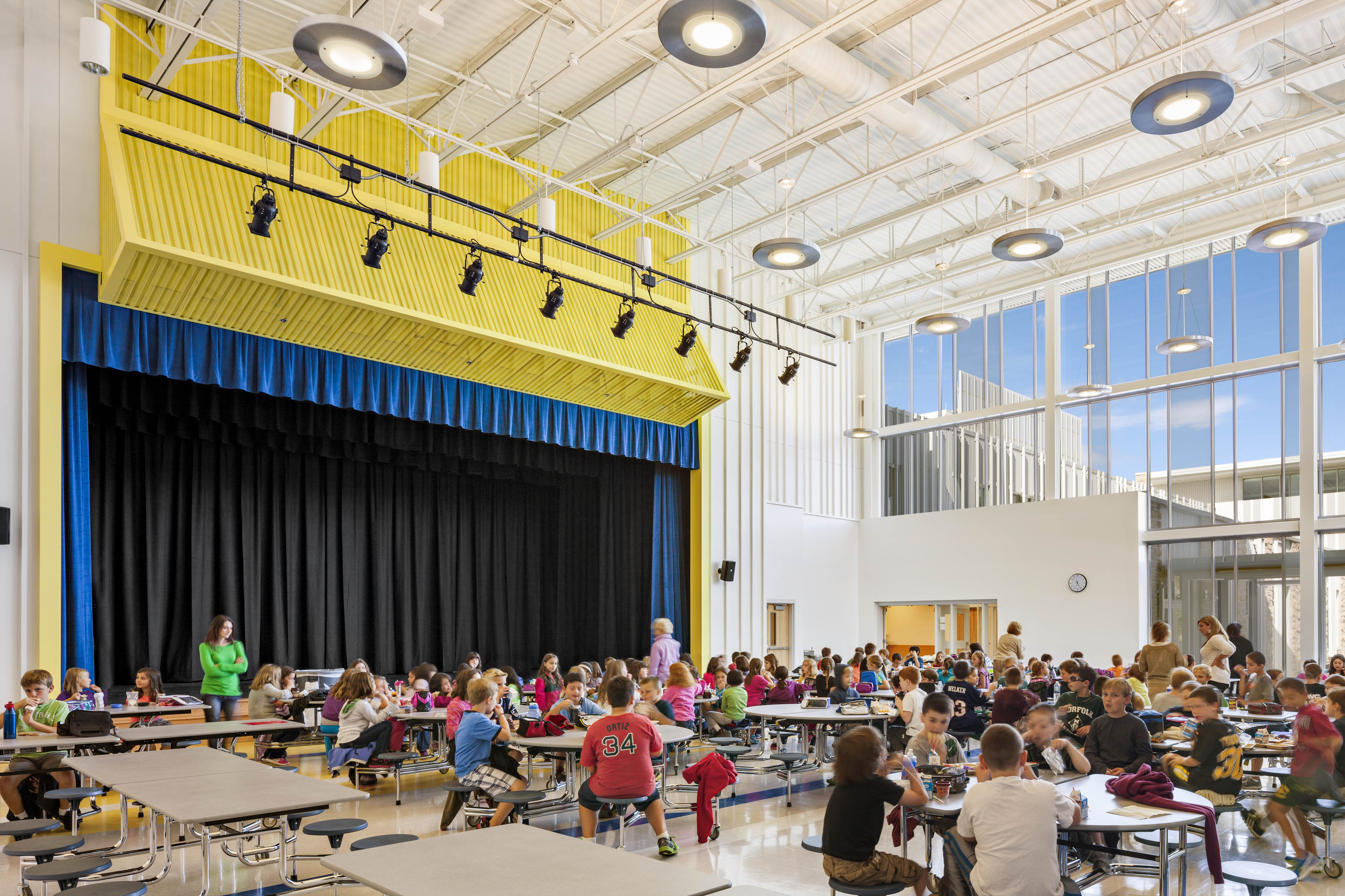 Students sat at cafeteria with stage to the left with yellow accents and a blue curtain 
