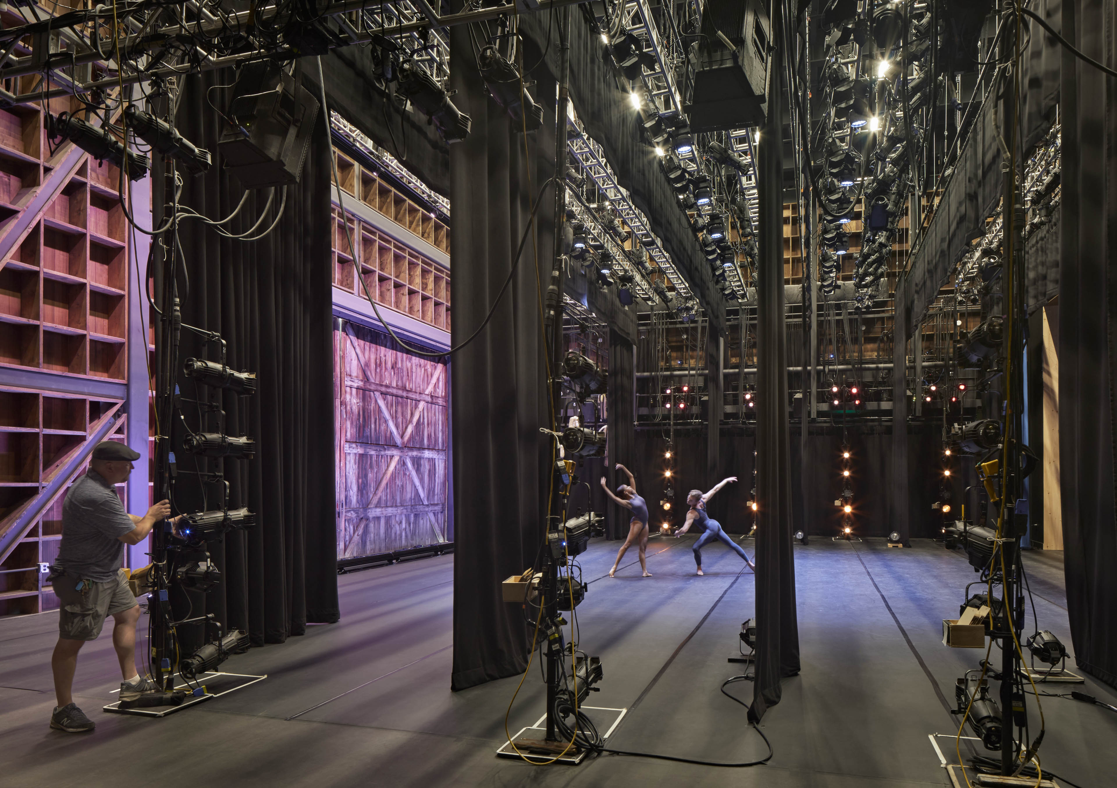 View from stage right, ballerinas on stage and wood in small square formation with barn door to the right and pink lighting displayed across backgrond