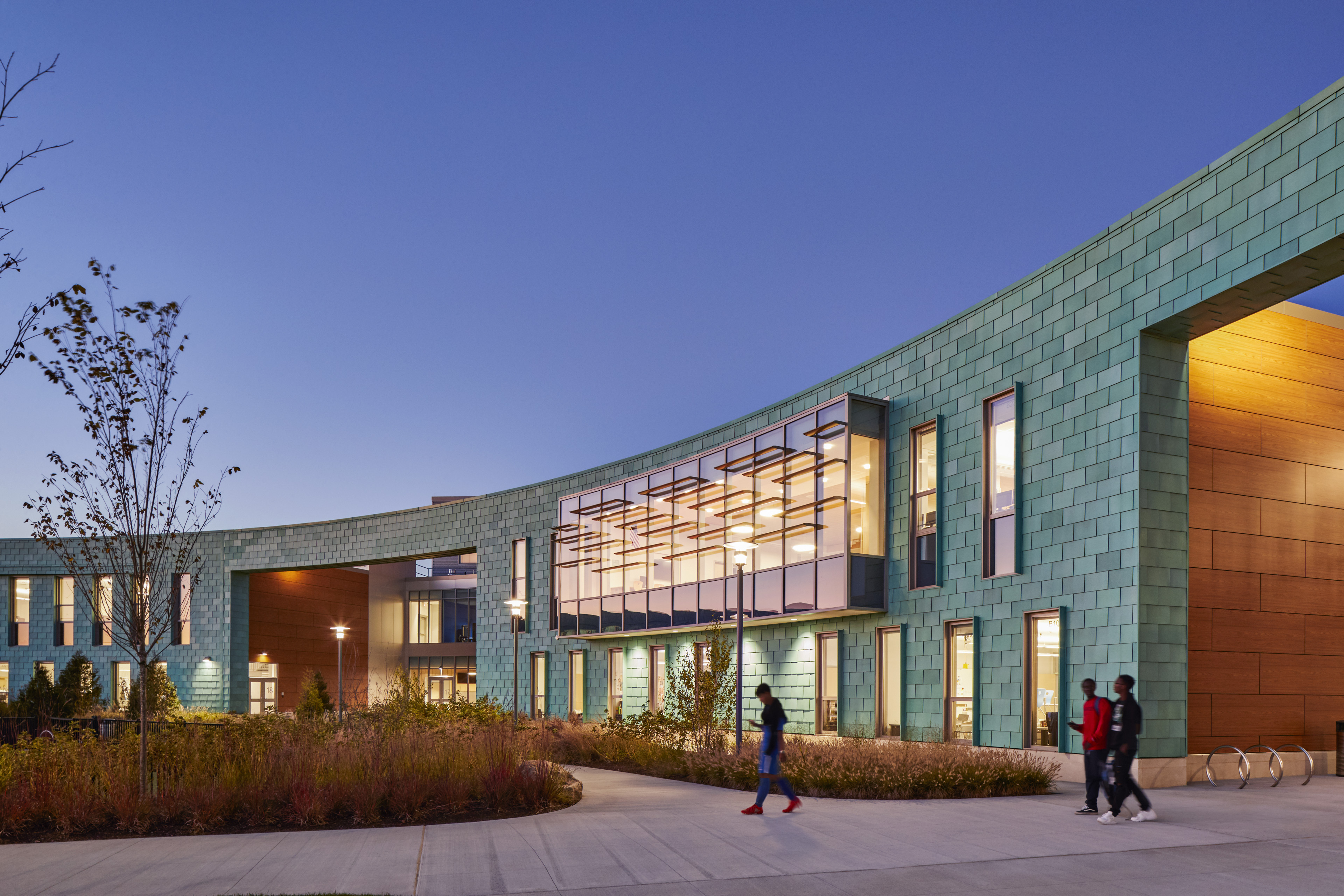 Exterior of school at dusk with aqua tiles and a curved facade, with large entrances to left and right with amber lights. Vertical windows in middle and foliage in front and blue sky