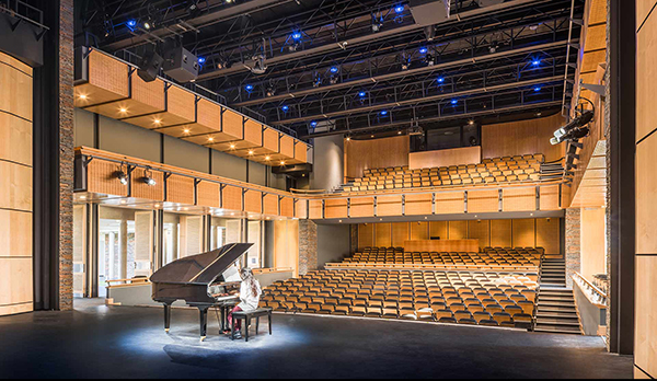 Theater from back of stage, girl sitting at grand piano with spotlight looking out at empty seats with lights on