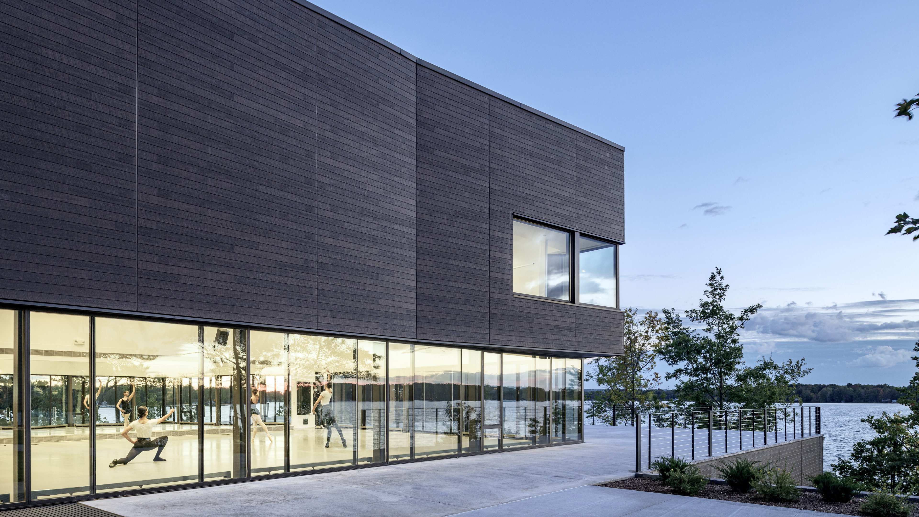 Yellow glow from ballet studio with ballerinas on first floor with glass windows and wood paneling on second floor. Overlooking water at dusk