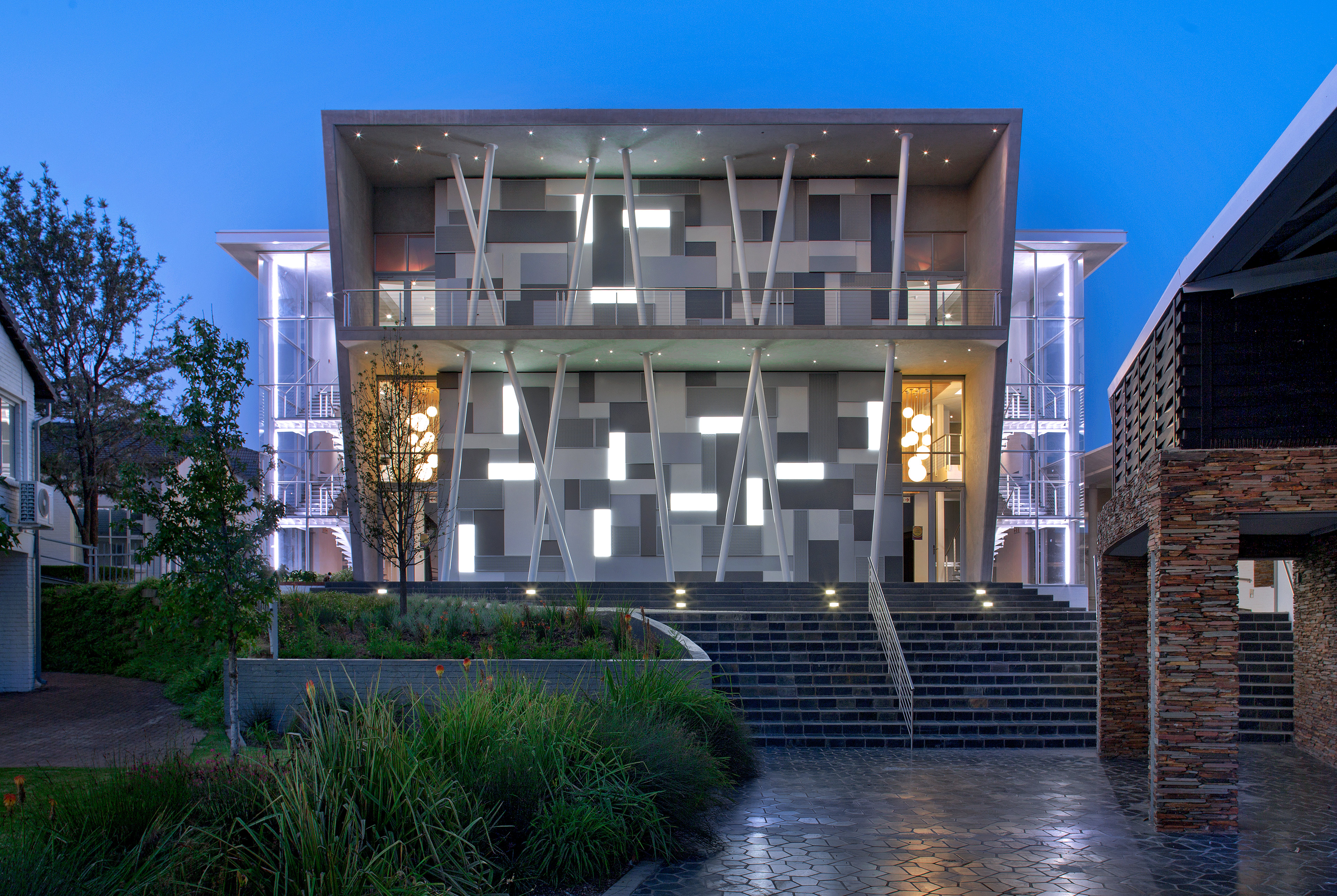Front entrance of building at dusk, with stairs leading up and poles criss-crossing in front. Building behind lit up and extending past on left and right side