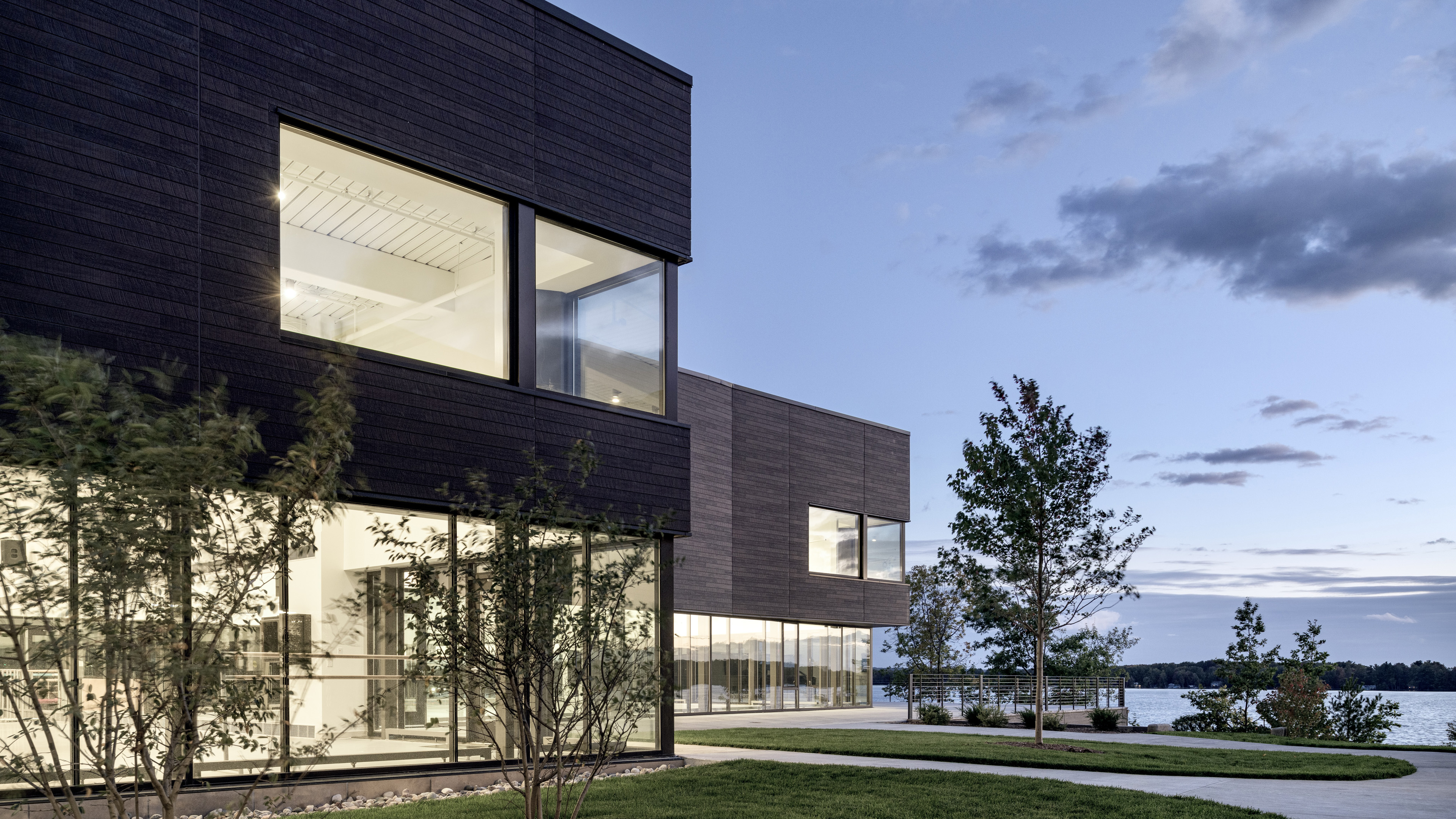 Exterior from courtyard at dusk with first floor all glass windows and second floor wood paneling overlooking trees and water