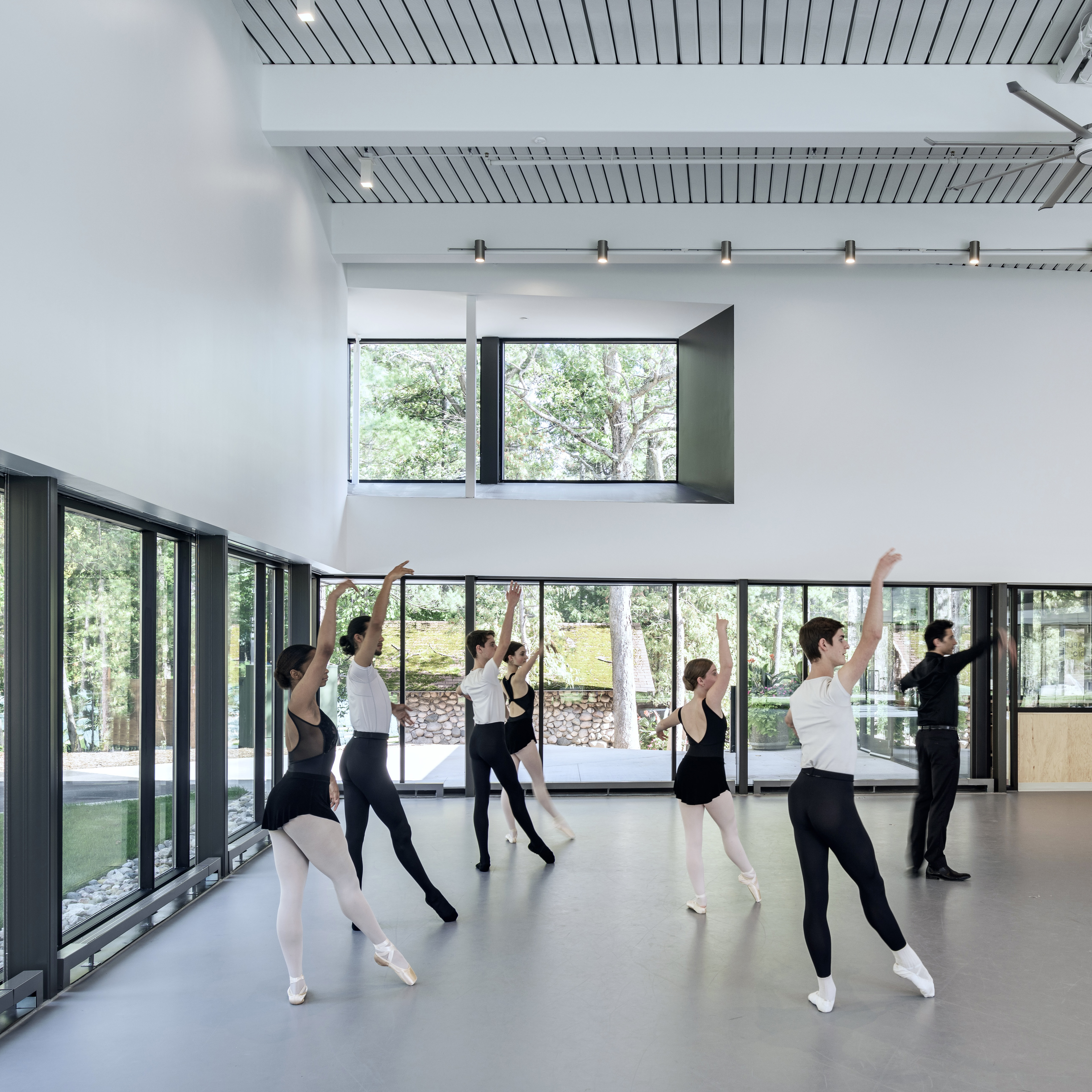 Interior of dance studio with ballerinas practicing on first floor surrounded by glass paneling, window of second floor looking out at trees