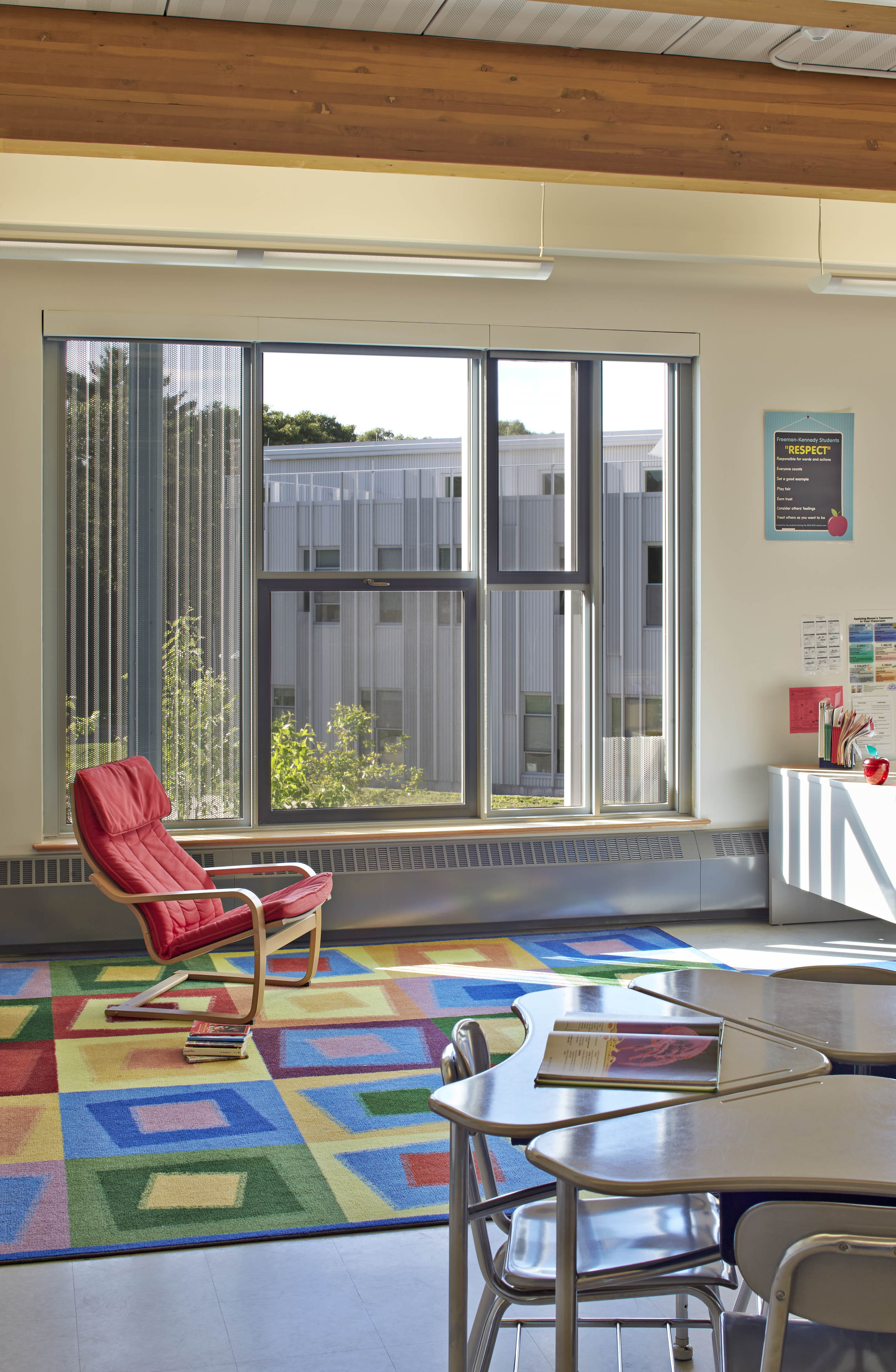 Red lounge seat on colorful carpet in corner of classroom with windows behind and desks to the right