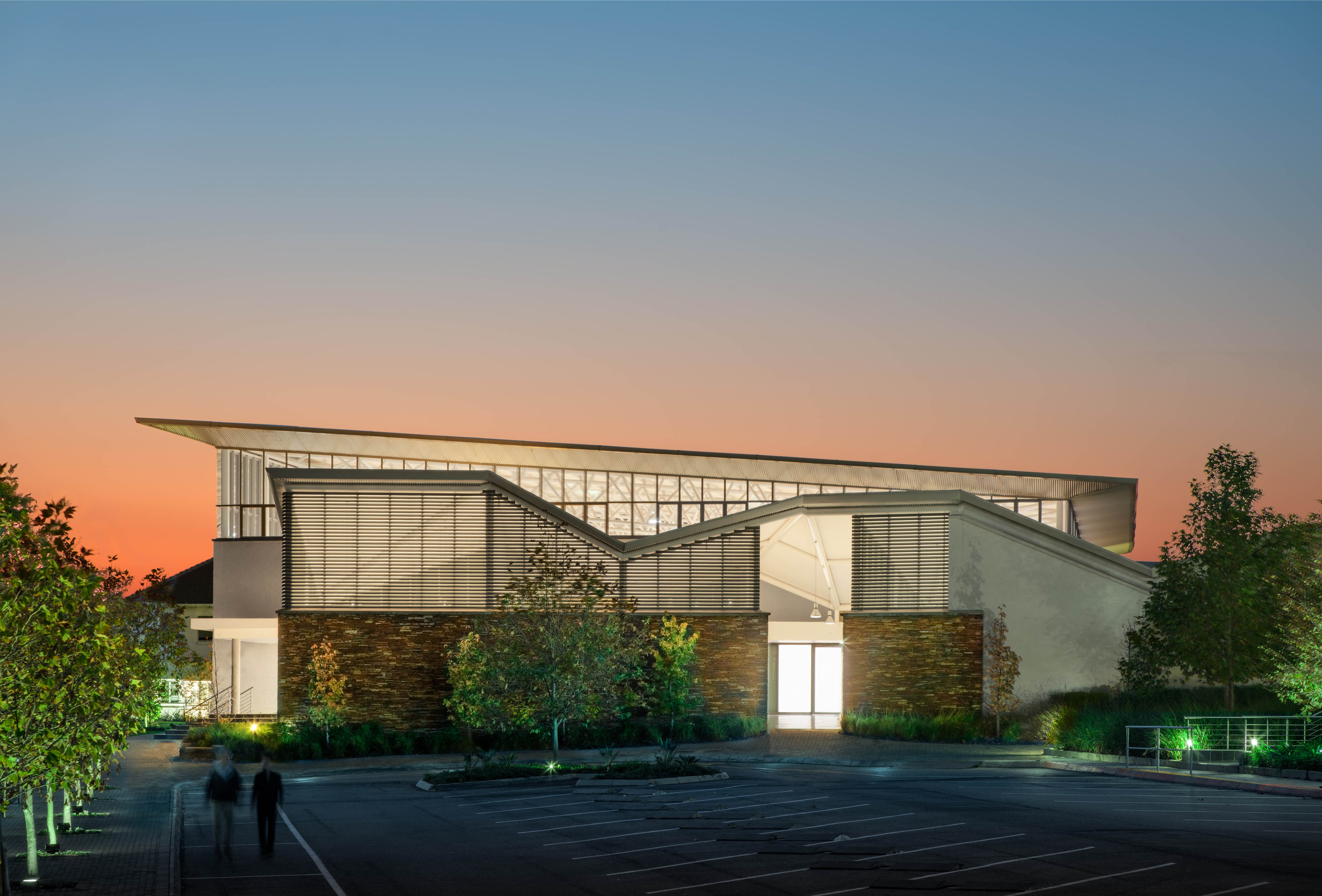 Exterior of building at night with orange to blue sunset behind, light emitting windows and slatted wood, parking lot in front