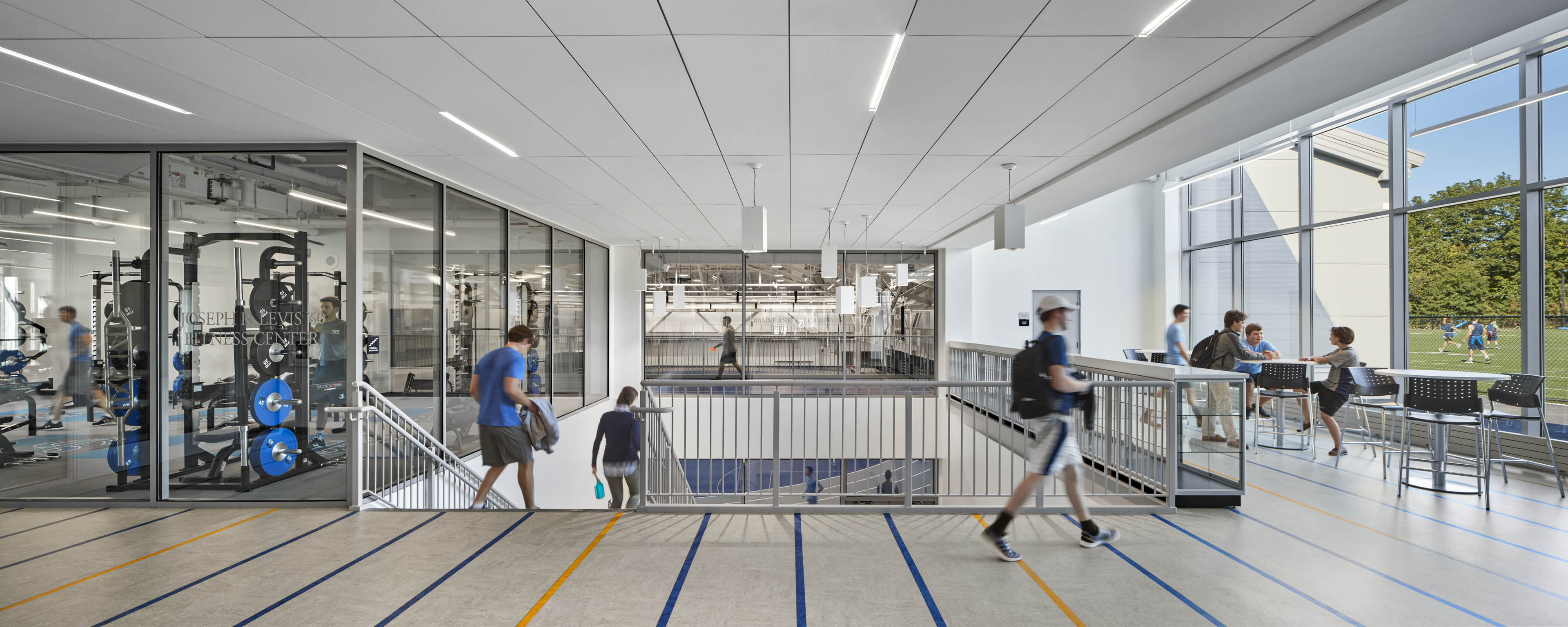 Students walking through corridor to staircase with workout room to left, and field with green grass to the windows to the right