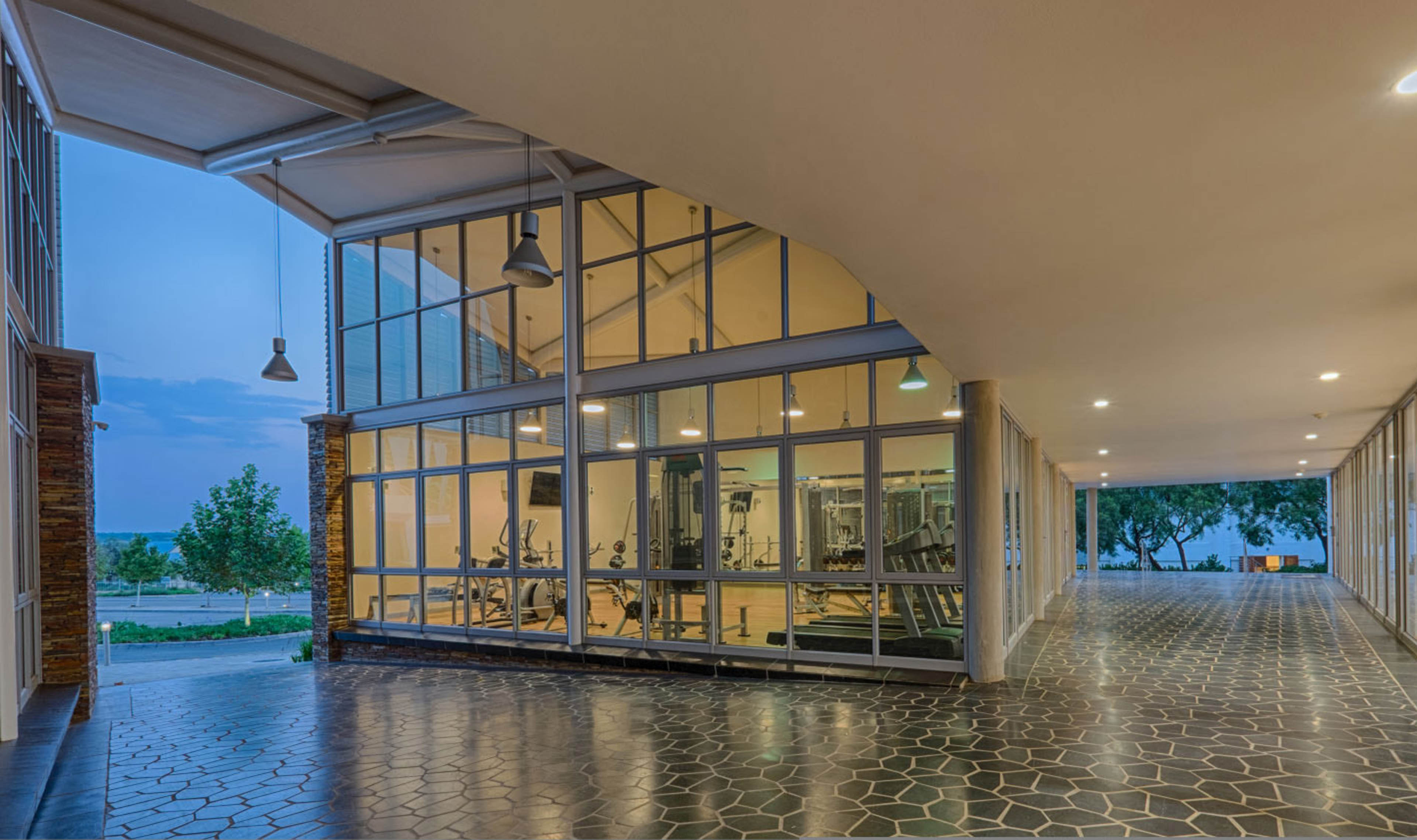 Exterior covered corridor at dusk with stone tiling looking at weight room in middle and trees with sky to left and right