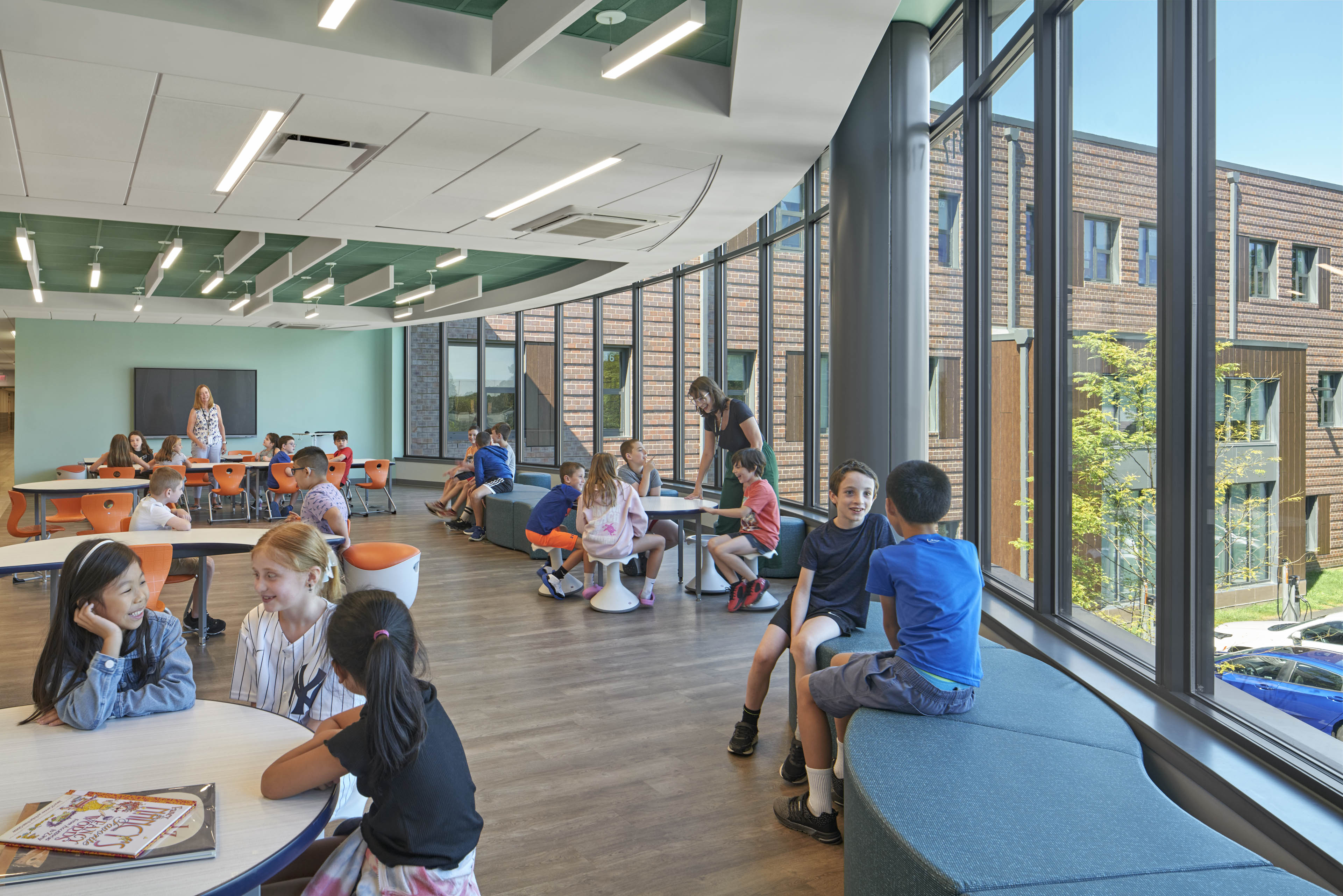 Students sitting around desks and lounge chairs with long vertical windows on right side looking out to courtyard