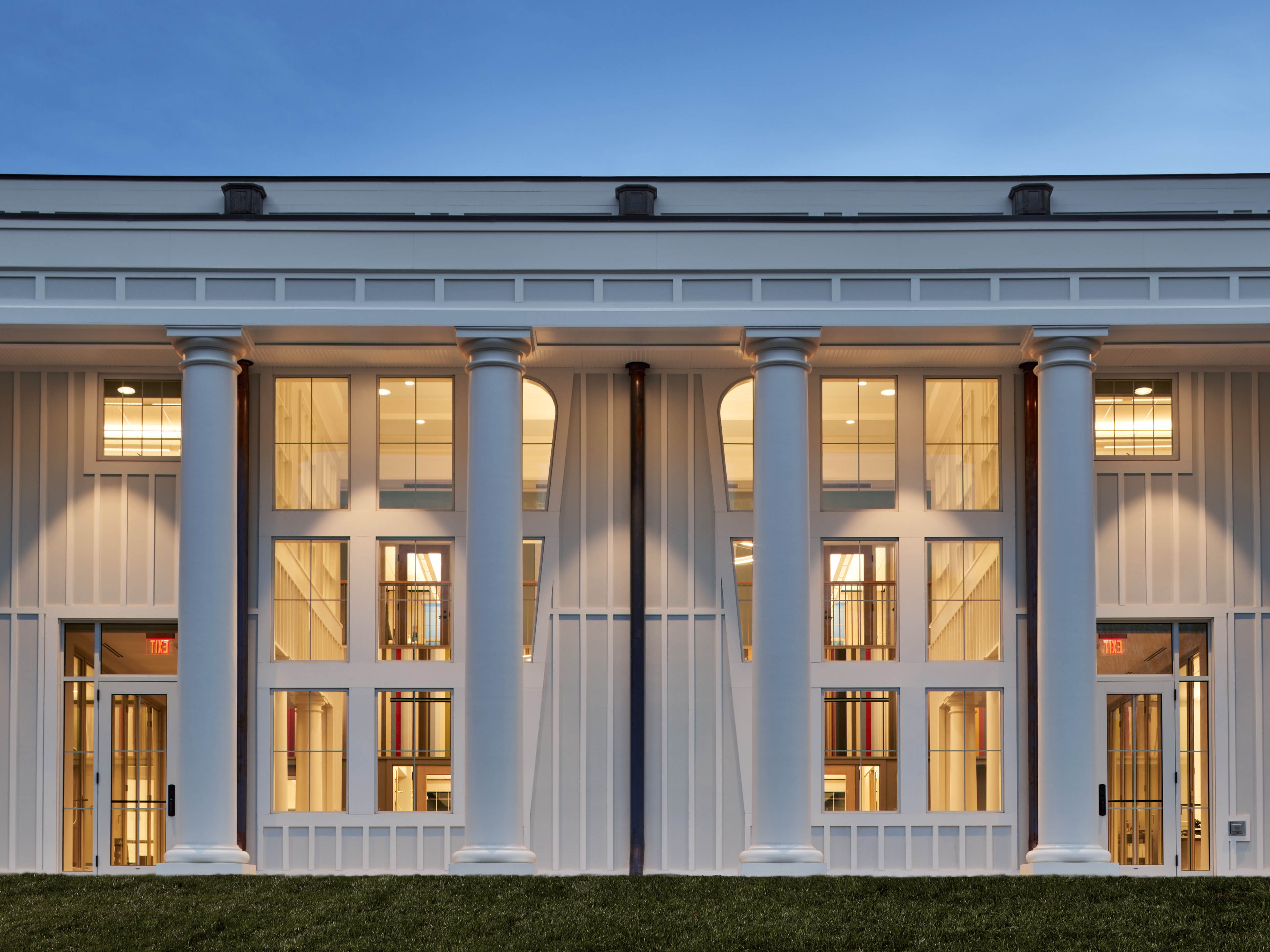 View of exterior at dusk with large white columns and warm yellow light escaping from windows, blue-purple sky above