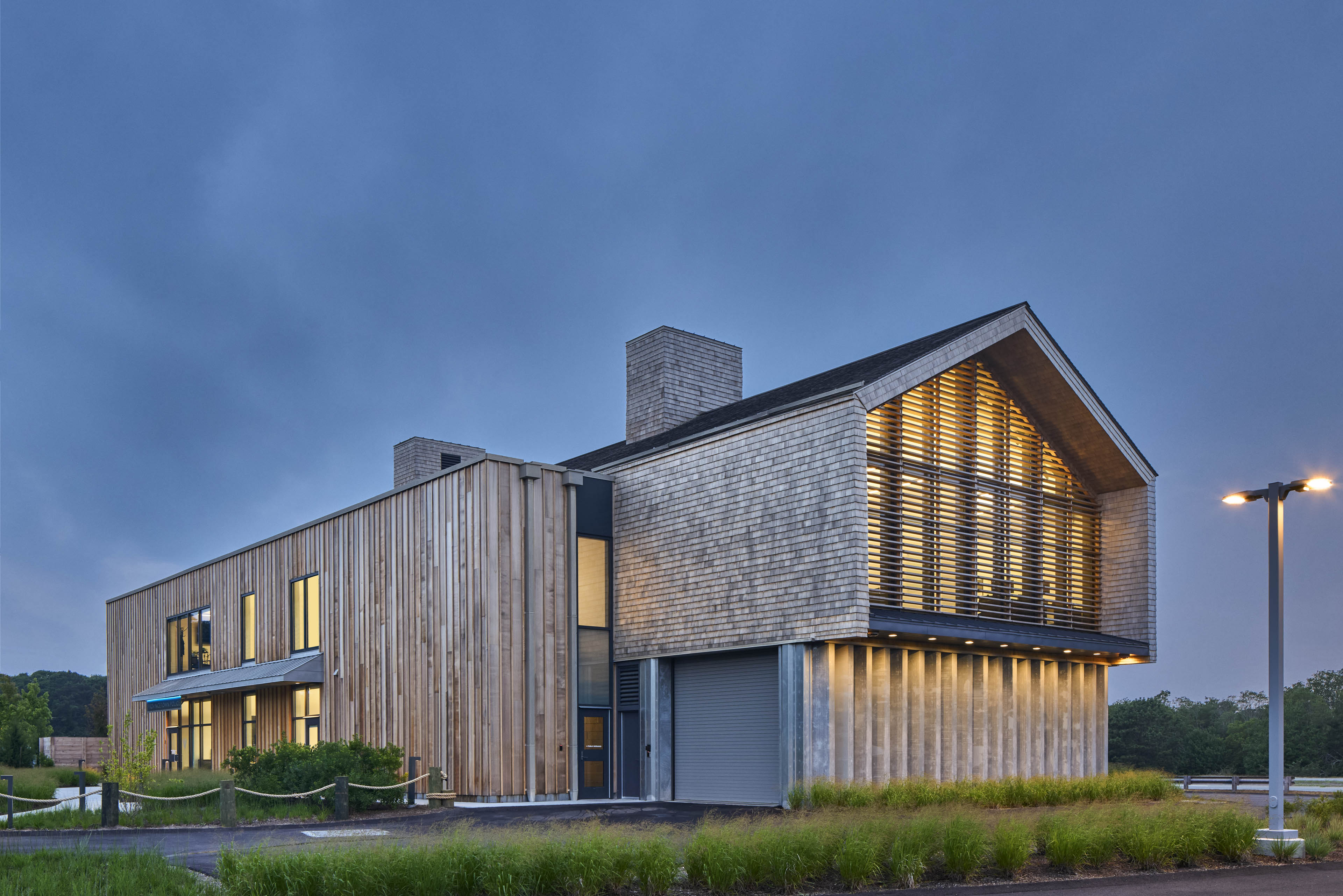 Side angle of police station at night, with natural greenery surrounding and a deep blue/grey sky, and yellow light glowing from within