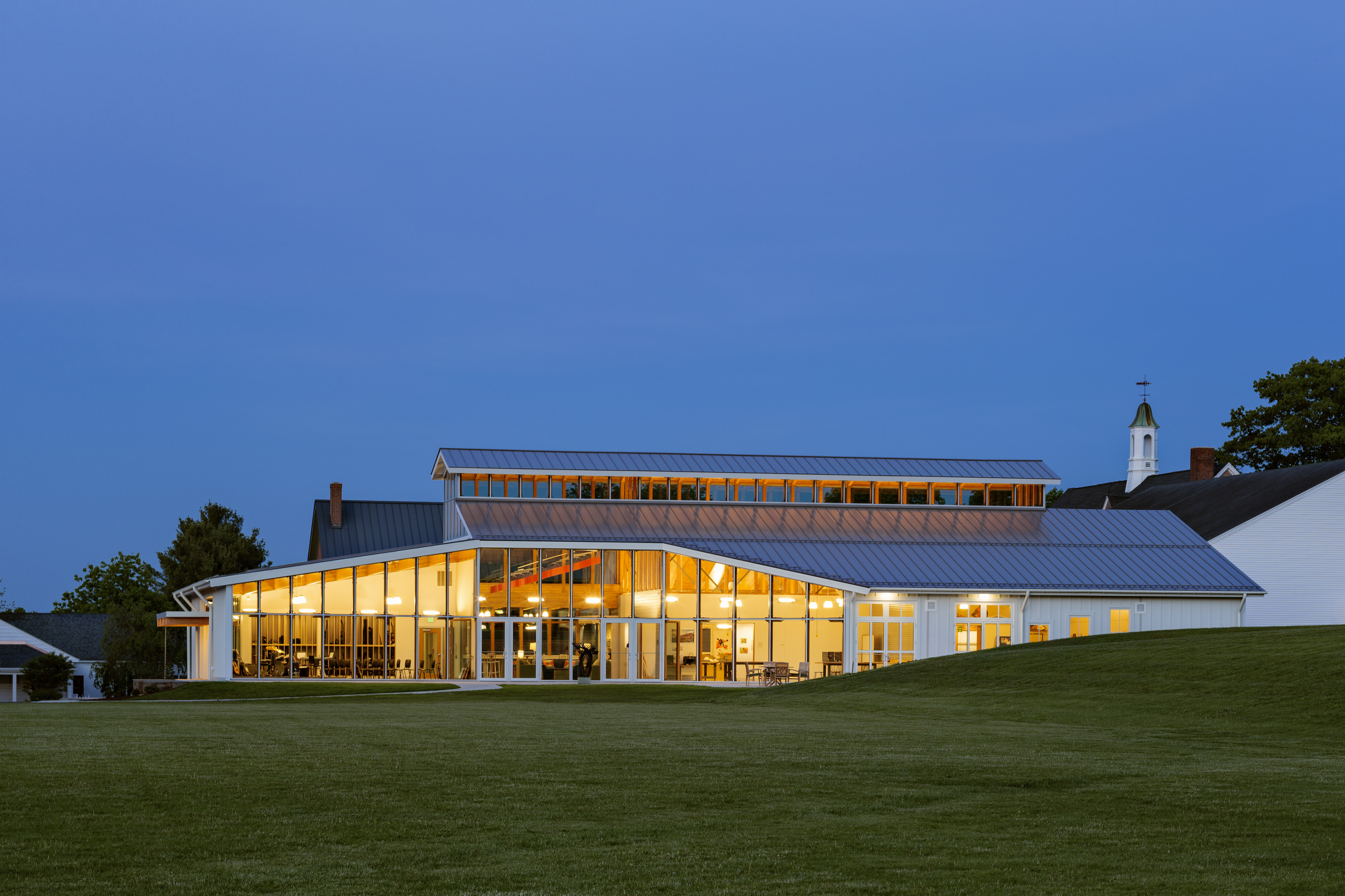 View at night of school from grassy hill, with long vertical windows with yellow orange light on building. Dark blue sky behind