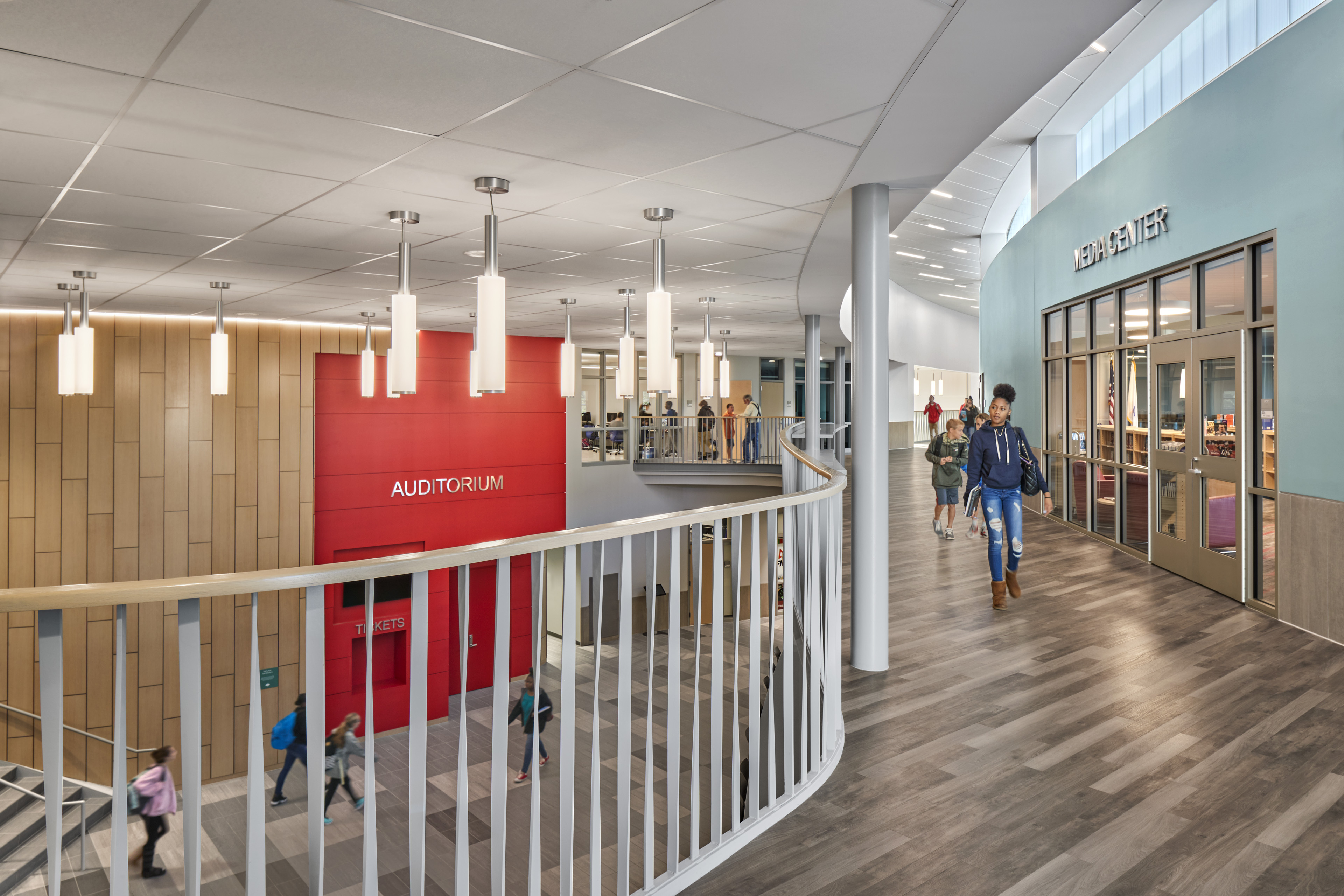 Hallway with wood floors and light blue media center to the right and students walking. Red auditorium and students walking to the left