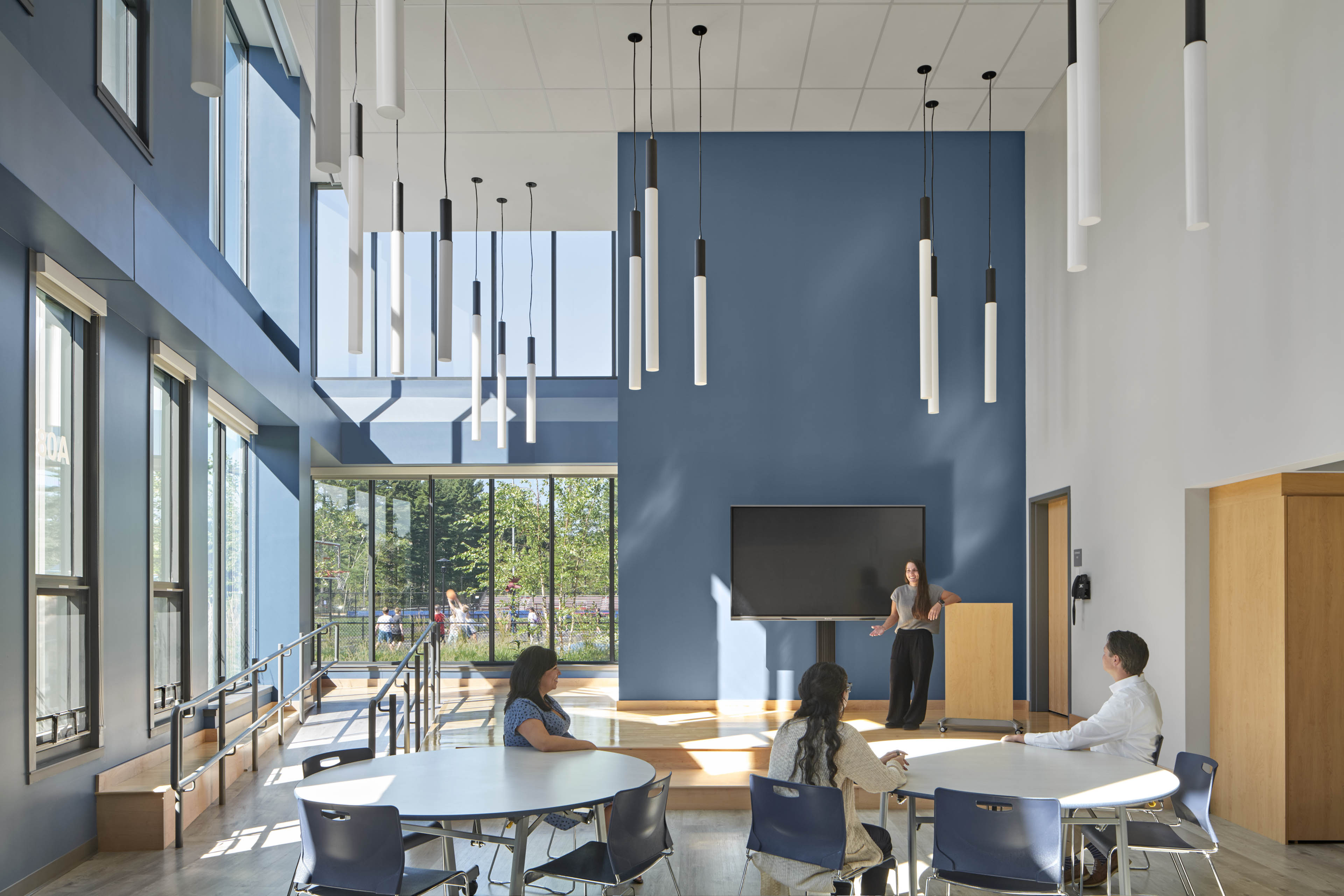 Presenter on stage with circular tables below, blue wall accents and window in back looking out to playground. Lights hanging down from ceiling 