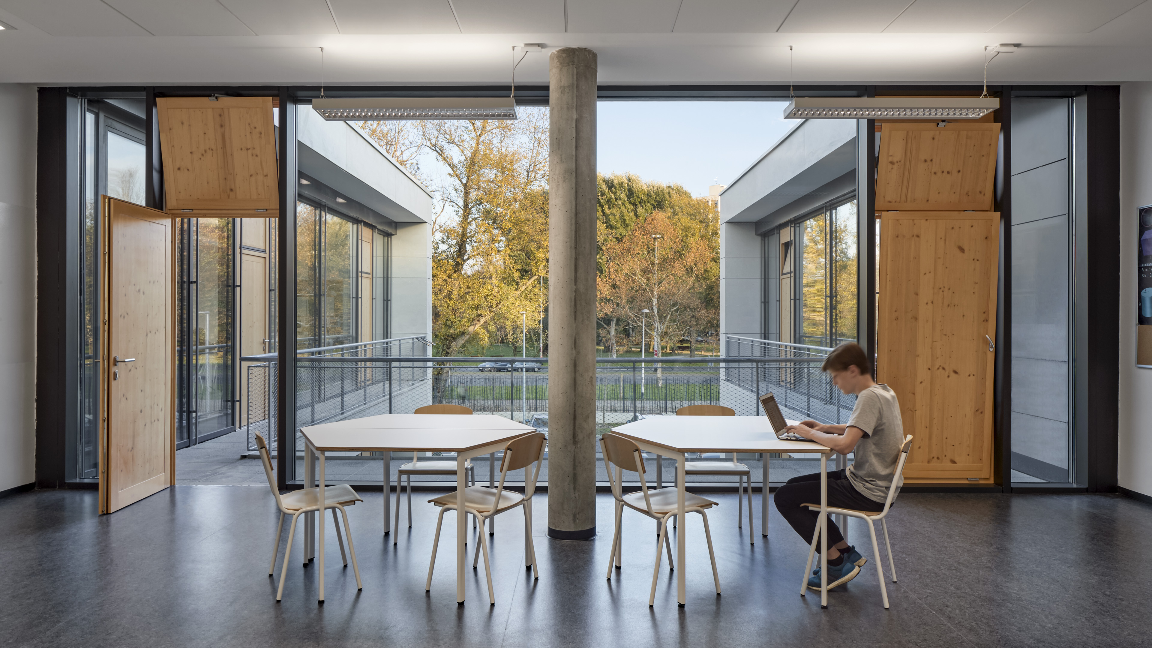Student seated at workspace with tables and chairs American International School of Zagreb looking over cluster of trees with a light wood door on either side