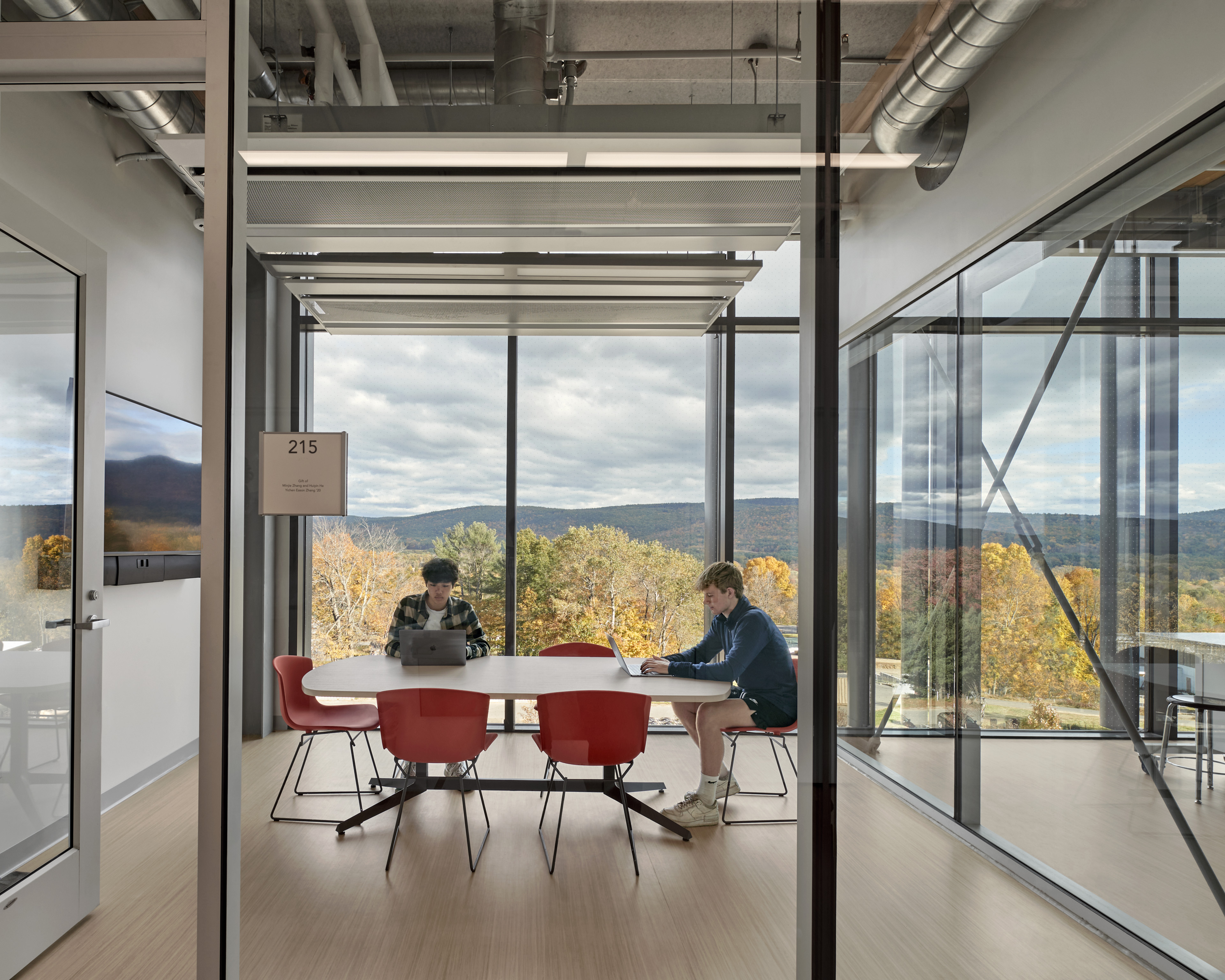 Students working at computer in breakout room with windows overlooking orange and yellow trees. 