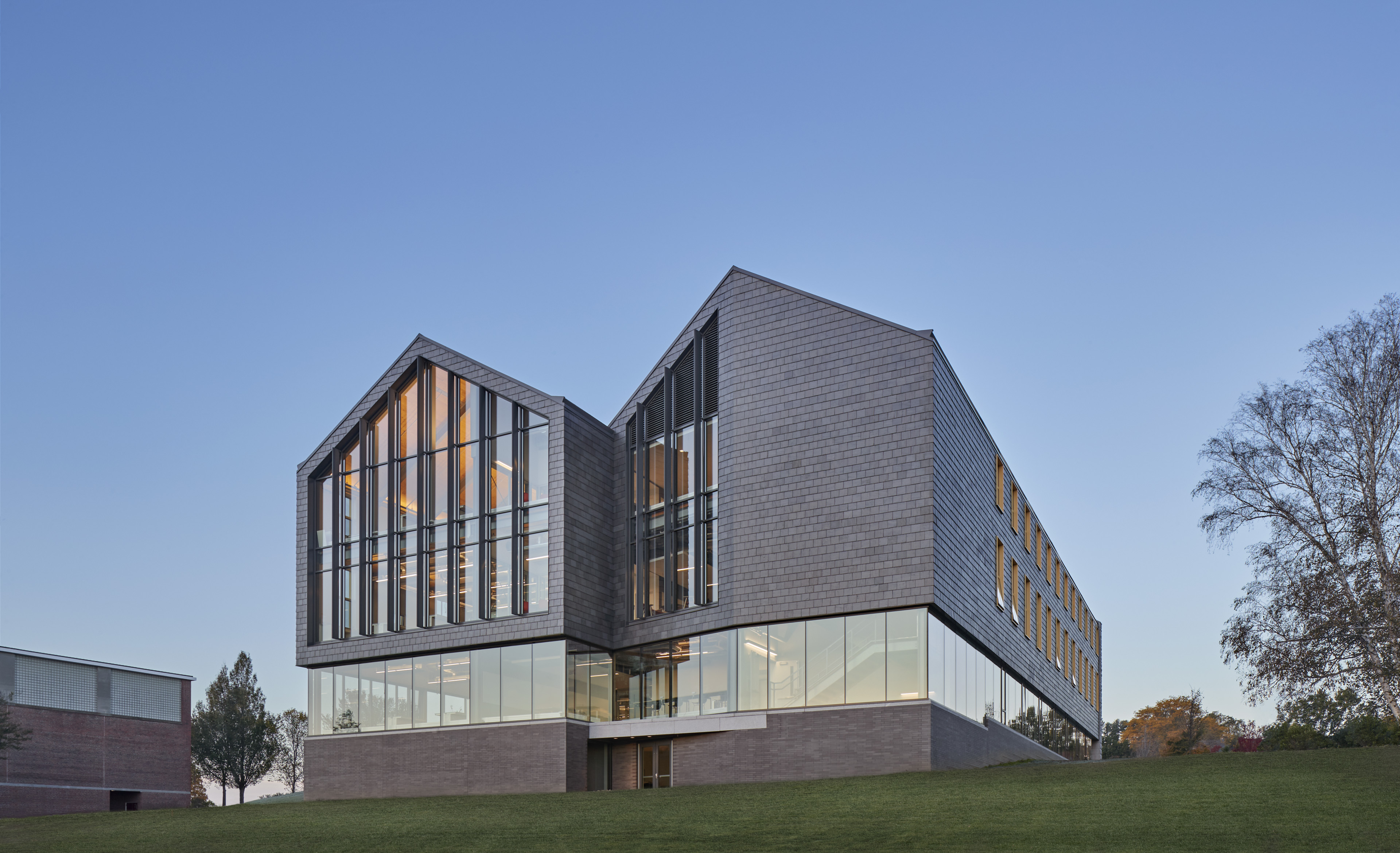 Side of building with a base of stone tiling, large vertical glass windows on first floor, and stone tiling encasing vertical windows until both peaks. Set on green grass and trees on both sides