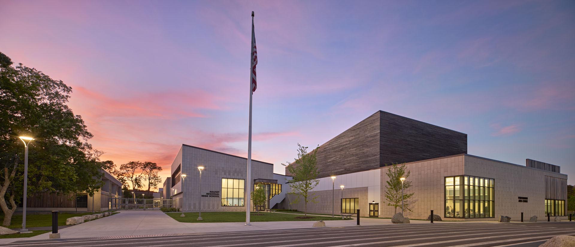 Entrance of school with pink and purple sunset draping the sky. Flagpole in center of image with amber glow from classrooms