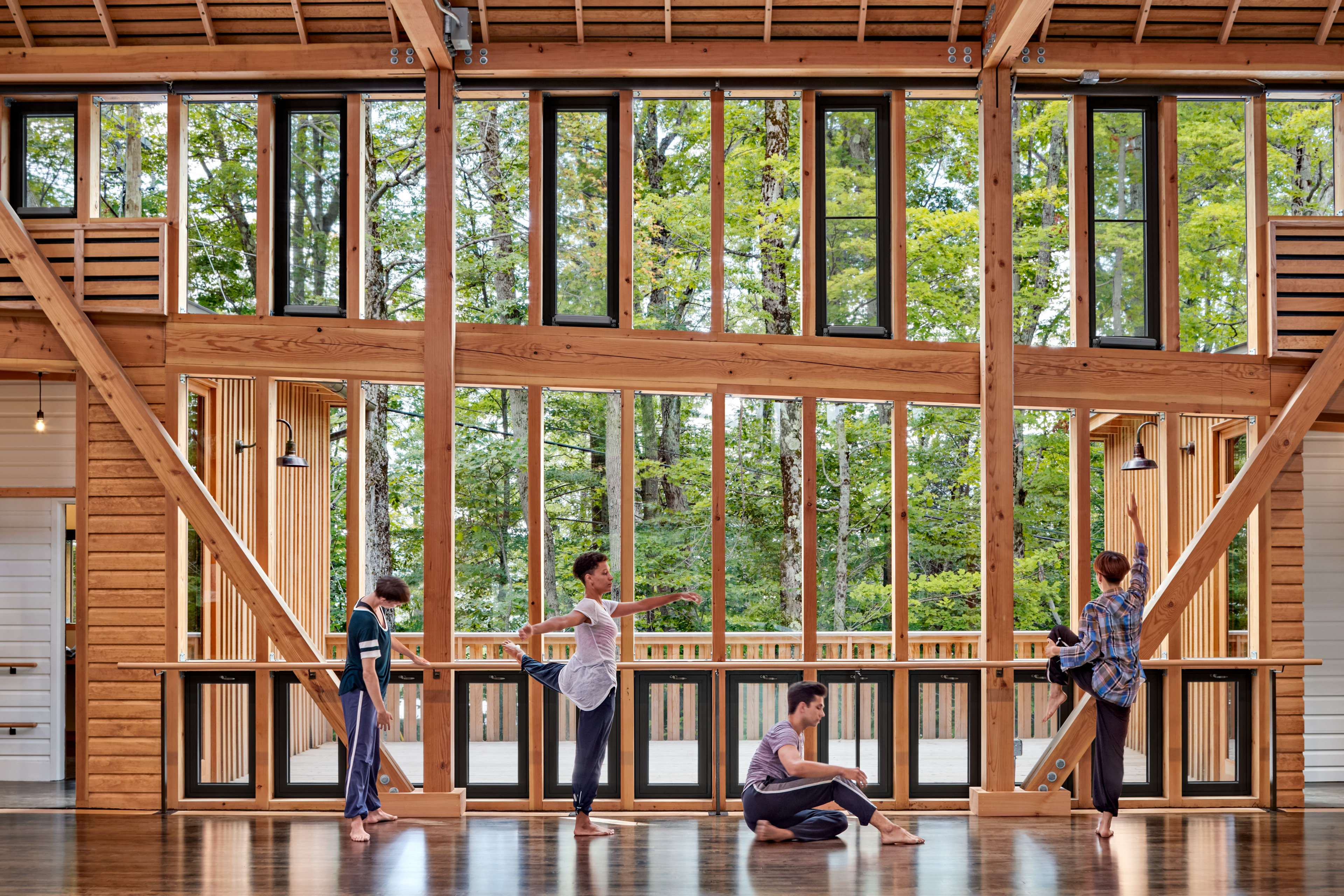 Ballerinas stretching at barre in dance studio with windows behind looking out at forest 