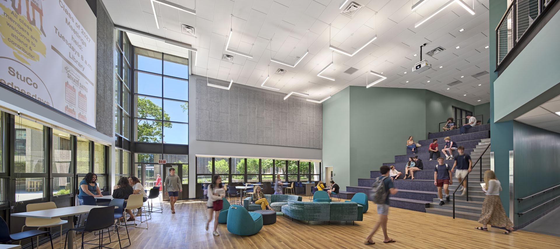 Student commons with lounge seats and stadium seating with green walls to the right. Large windows on left side looking out at foliage