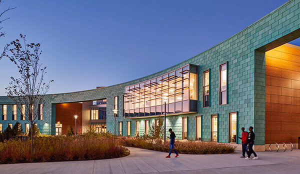 Exterior of school at dusk with aqua tiles and a curved facade, with large entrances to left and right with amber lights. Vertical windows in middle and foliage in front and blue sky