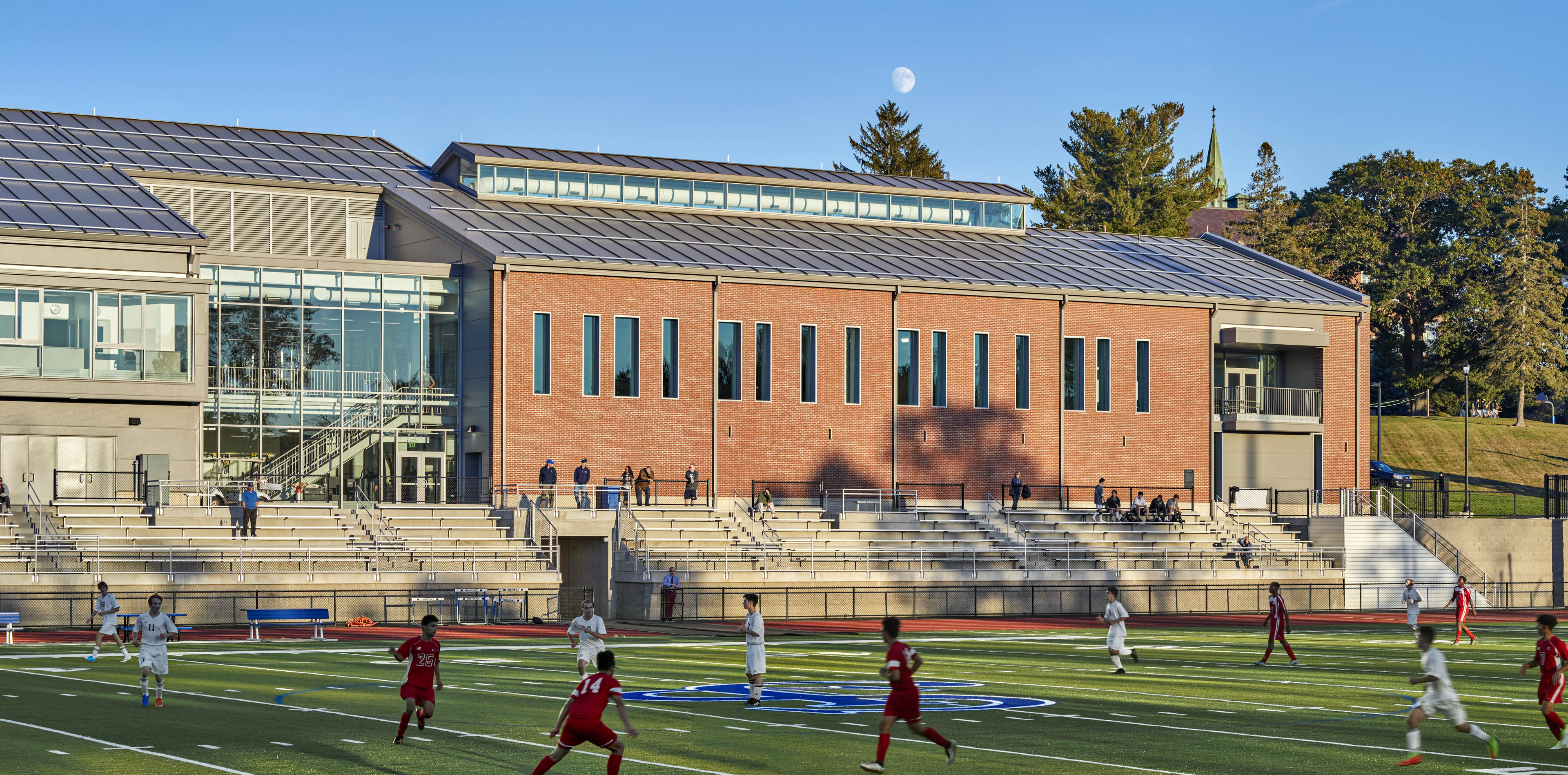 Side angle view of exterior of school with students practicing on football field, stadium seating behind and light shining on building
