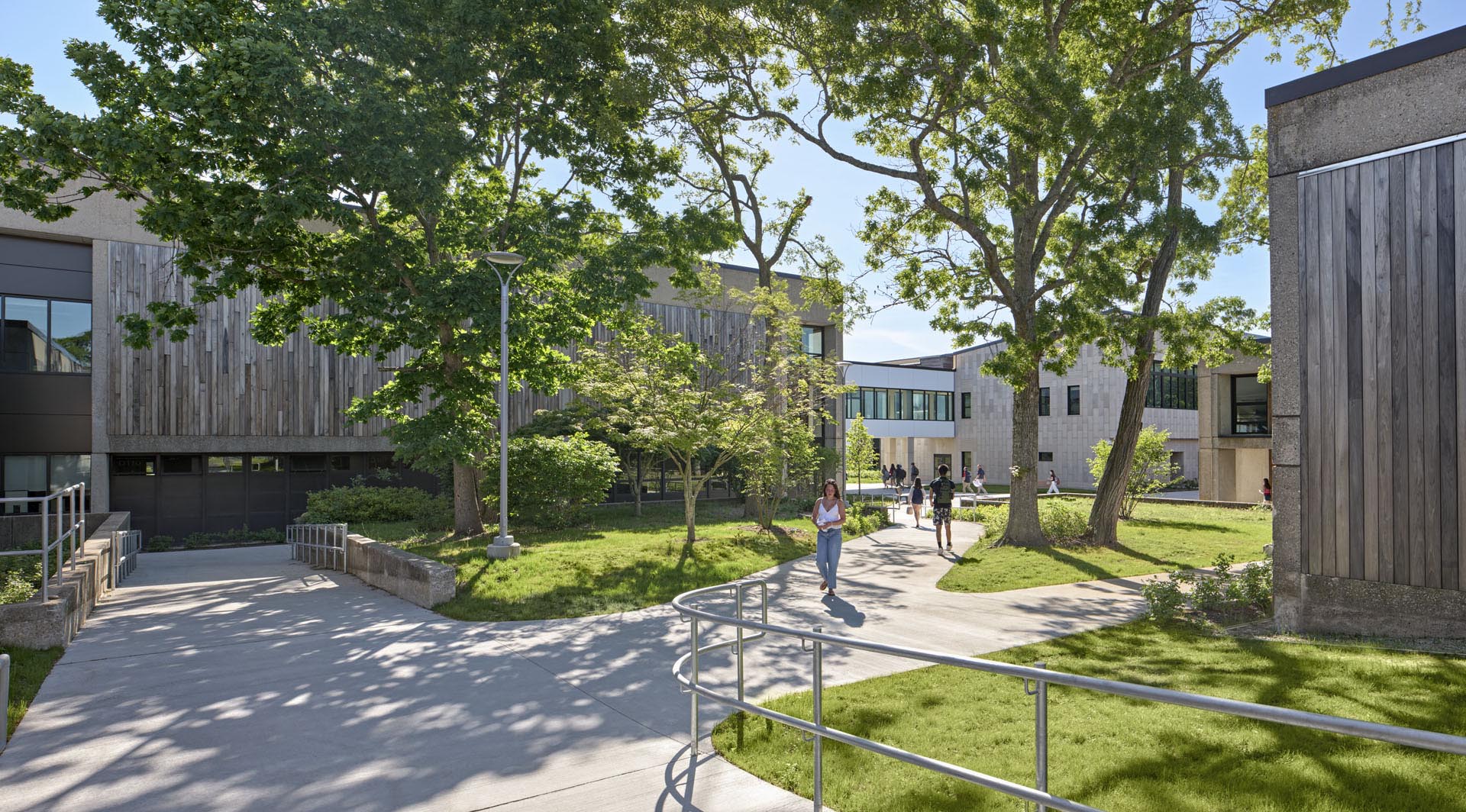 Courtyard with tall tree coverage, green grass and students on walkways. Wood paneled buildings to left and right, with skybridge connection in back 